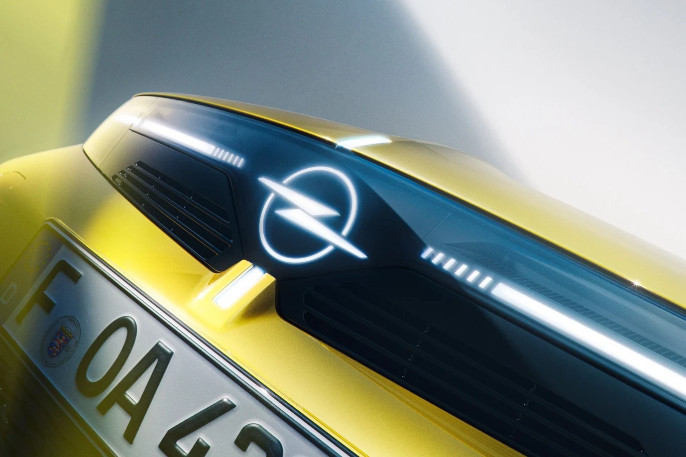 Nowy Opel Astra został wyposażony w nowy przód z nowym, podświetlanym logo marki. Przód nowoczesnego, żółtego samochodu marki Opel z podświetlonym logotypem na czarnej listwie oraz fragmentem tablicy rejestracyjnej.