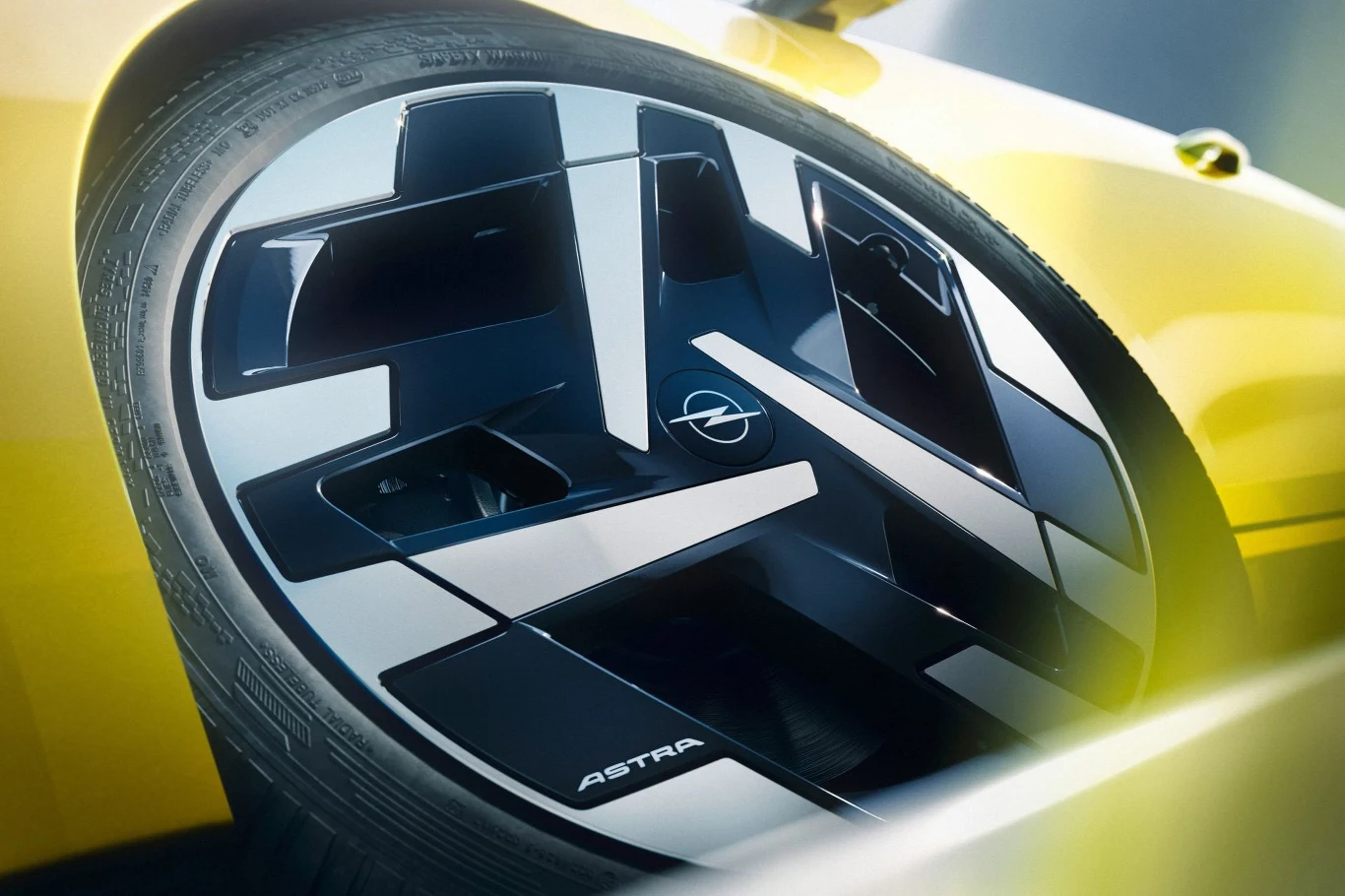 Opel Astra po modernizacji został wyposażony w nowe wzory 17- i 18-calowe felg. Nowoczesna felga samochodowa z charakterystycznym designem, widoczny emblemat marki Opel oraz nazwa modelu Astra na obręczy, żółty lakier karoserii w tle.