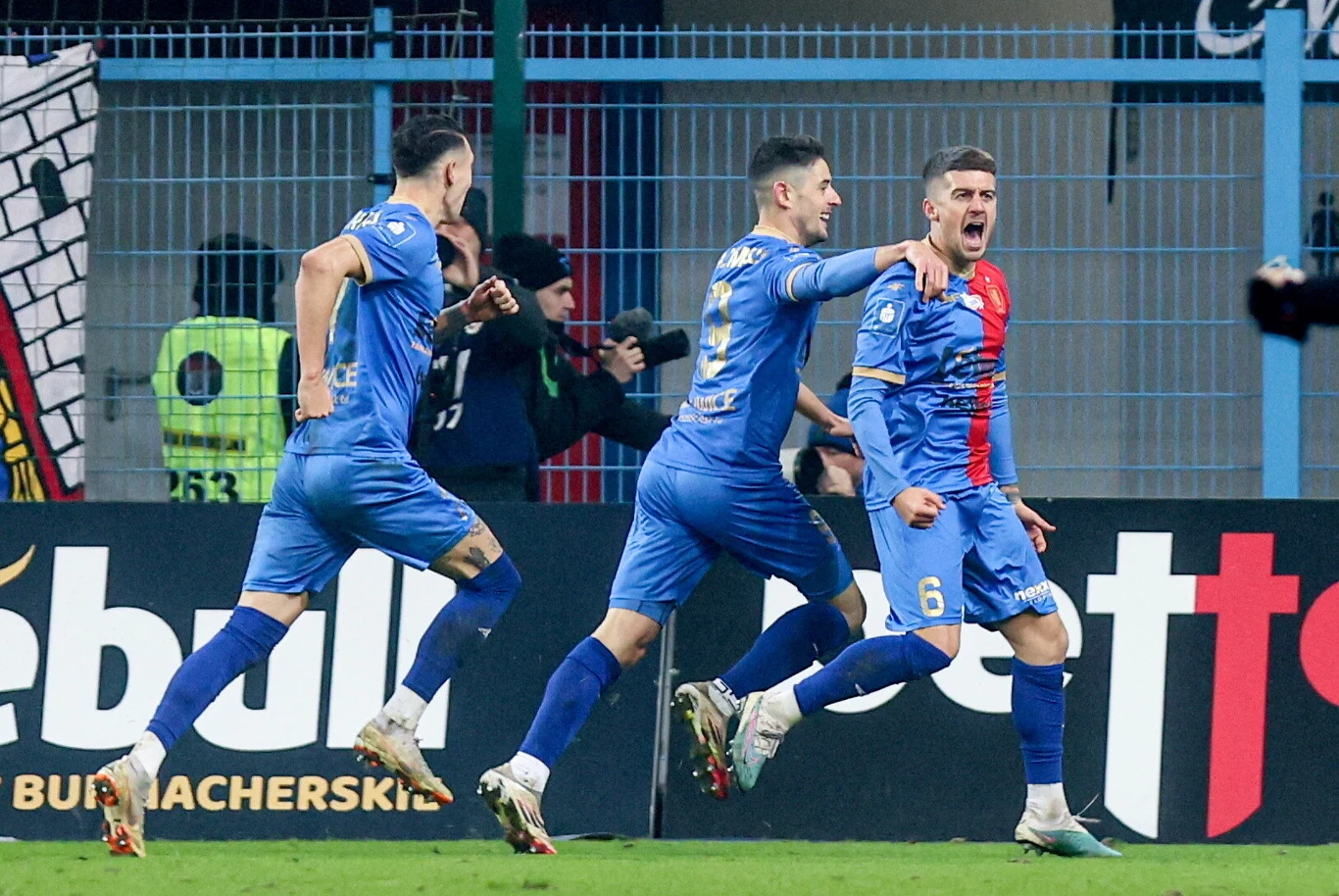 Piłkarze w niebieskich strojach świętują zdobycie gola na tle trybun i zabezpieczeń stadionowych. Jeden z zawodników wyraża emocje poprzez krzyk i uniesione ręce, pozostali biegną za nim z radością.