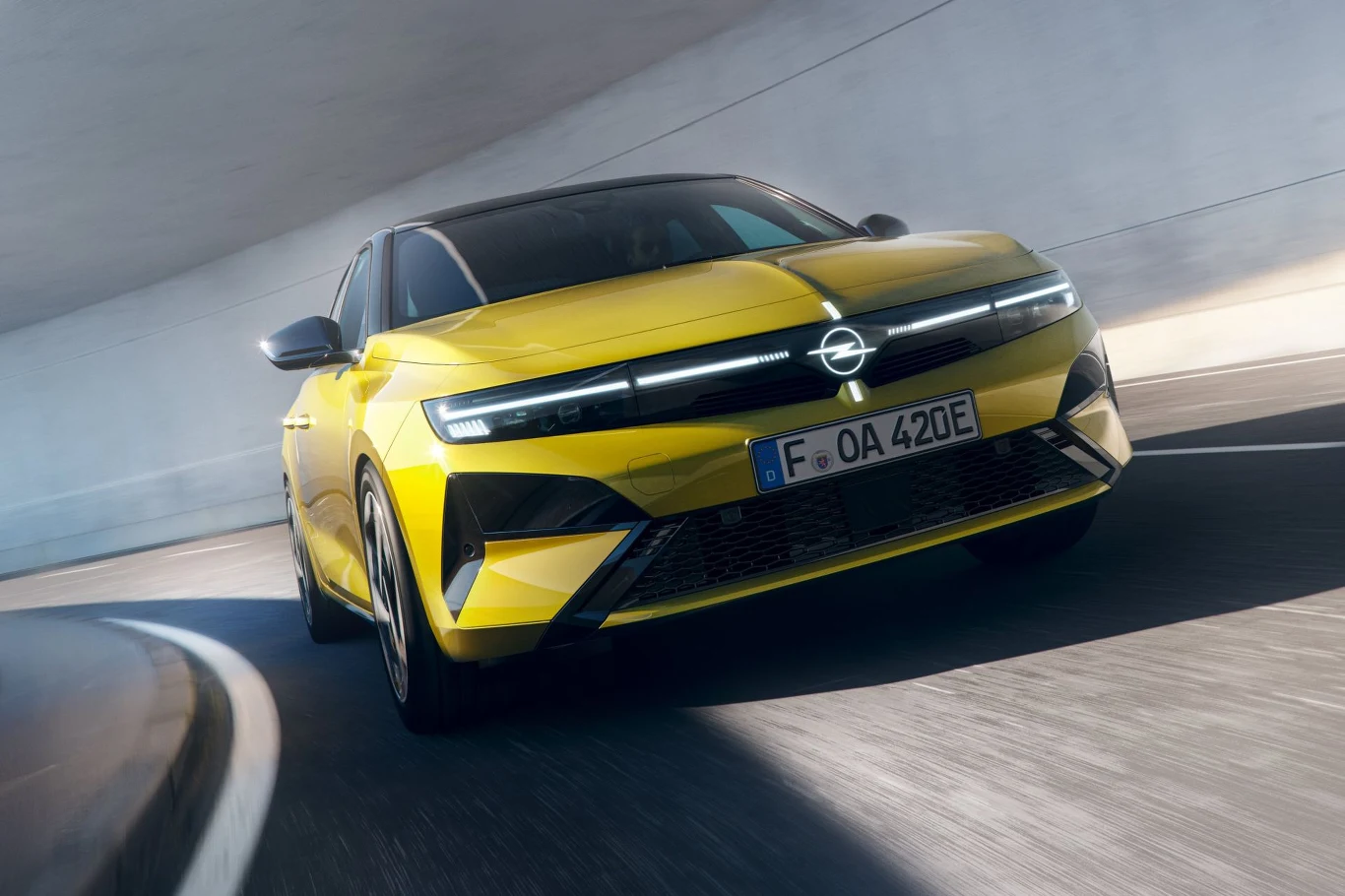 Opel Astra po liftingu został wyposażony w nieco większy akumulator, dzięki czemu przejedzie na jednym ładowaniu do 454 km. Jaskrawożółty samochód marki Opel porusza się dynamicznie po łuku pod jasnym wiaduktem, na tablicy rejestracyjnej widoczny numer F OA 420E. Nowoczesna stylistyka samochodu podkreślona ostrymi liniami i ciemnymi szybami.