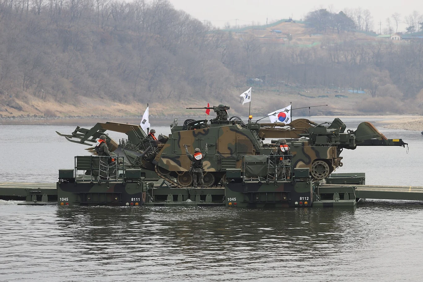Czołg saperski K600 Rhino transportowany przez rzekę podczas ćwiczeń Wojskowy pojazd opancerzony przeprawia się przez rzekę na specjalnej pontonowej platformie, wokół widoczne są żołnierze oraz flagi Korei Południowej. W tle widać pagórkowaty krajobraz porośnięty drzewami.