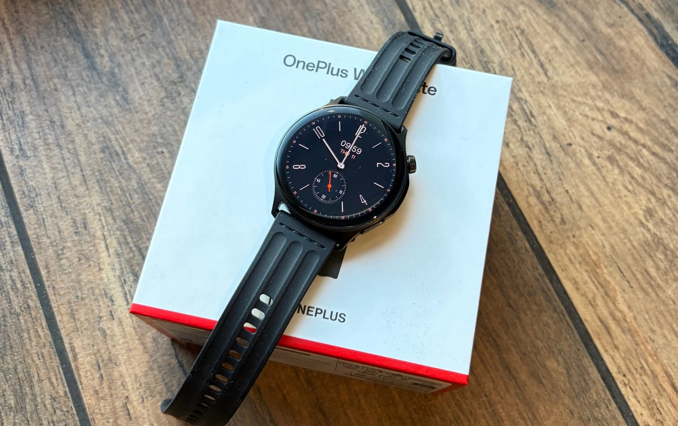 Test OnePlus Watch Lite. Tani smartwatch z wieloma funkcjami. Czarny smartwatch OnePlus leży na swoim oryginalnym białym opakowaniu z czerwonymi akcentami, całość ustawiona na drewnianej podłodze.