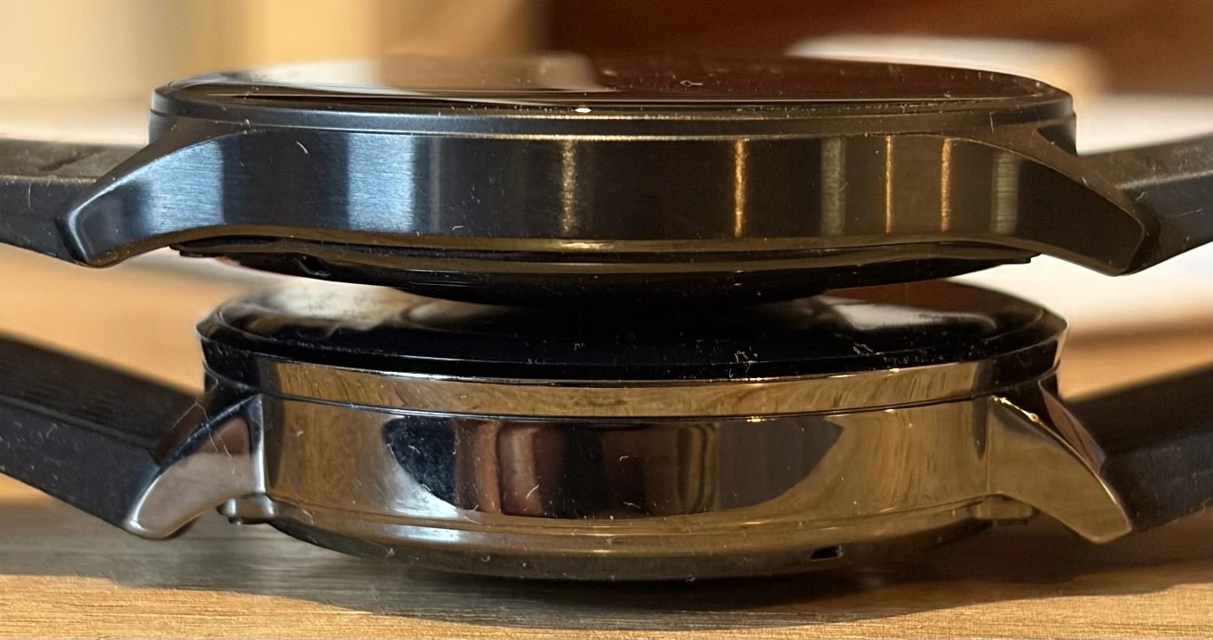 OnePlus Watch Lite jest cieńszy od Watcha 3 43 mm. Dwa inteligentne zegarki leżące jeden na drugim, widok z boku uwydatnia różnicę w grubości obudowy oraz wykończeniu metalu. Tło rozmyte, skupienie na detalach zegarków.