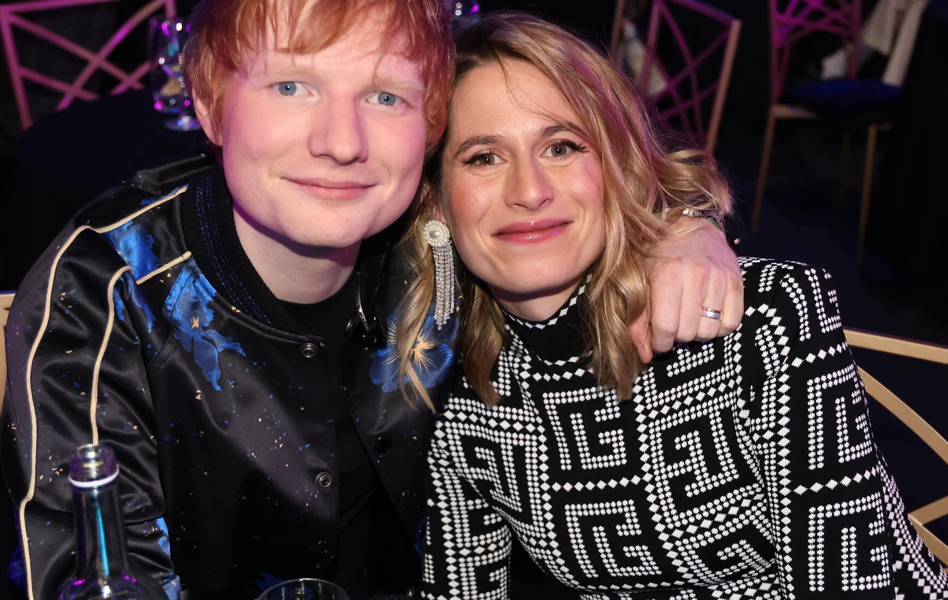 Ed Sheeran ze swoją partnerką - Cherry Seahorn Dwie osoby siedzą obok siebie, przytulone, patrzą prosto w obiektyw i uśmiechają się. W tle widoczne są rozmyte krzesła i stoły, panuje przyjazna atmosfera.
