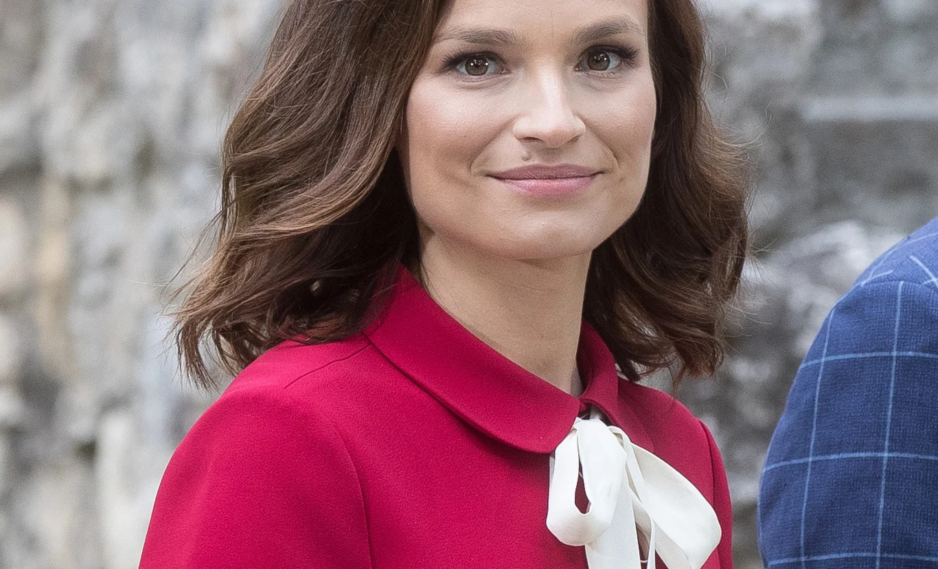 Młoda kobieta z półdługimi brązowymi włosami uśmiecha się delikatnie, ubrana w elegancką, czerwoną sukienkę z białą kokardą pod szyją, w tle rozmyte, kamienne ściany.