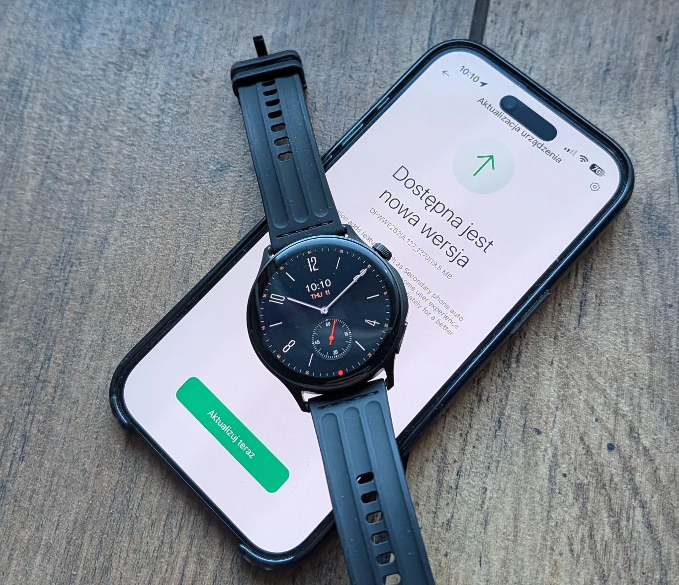 OnePlus Watch Lite to smartwatch, który współpracuje nie tylko z Androidem, ale też iPhone'ami. Smartfon z włączonym ekranem prezentującym komunikat o dostępnej nowej wersji oprogramowania, na ekranie położony czarny sportowy zegarek smartwatch z okrągłą tarczą i silikonowym paskiem, całość na drewnianym blacie.