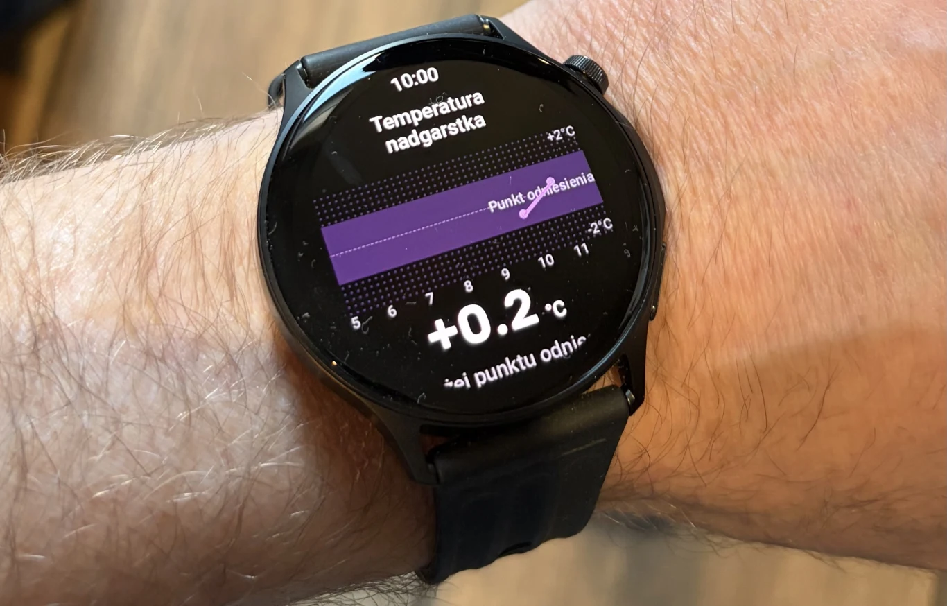 OnePlus Watch Lite pozwala monitorować temperaturę nadgarstka. Smartwatch na ręce mierzący temperaturę nadgarstka, na ekranie widoczny wykres oraz wynik pomiaru +0,2°C względem punktu odniesienia, pasek zegarka czarny, tło rozmyte.