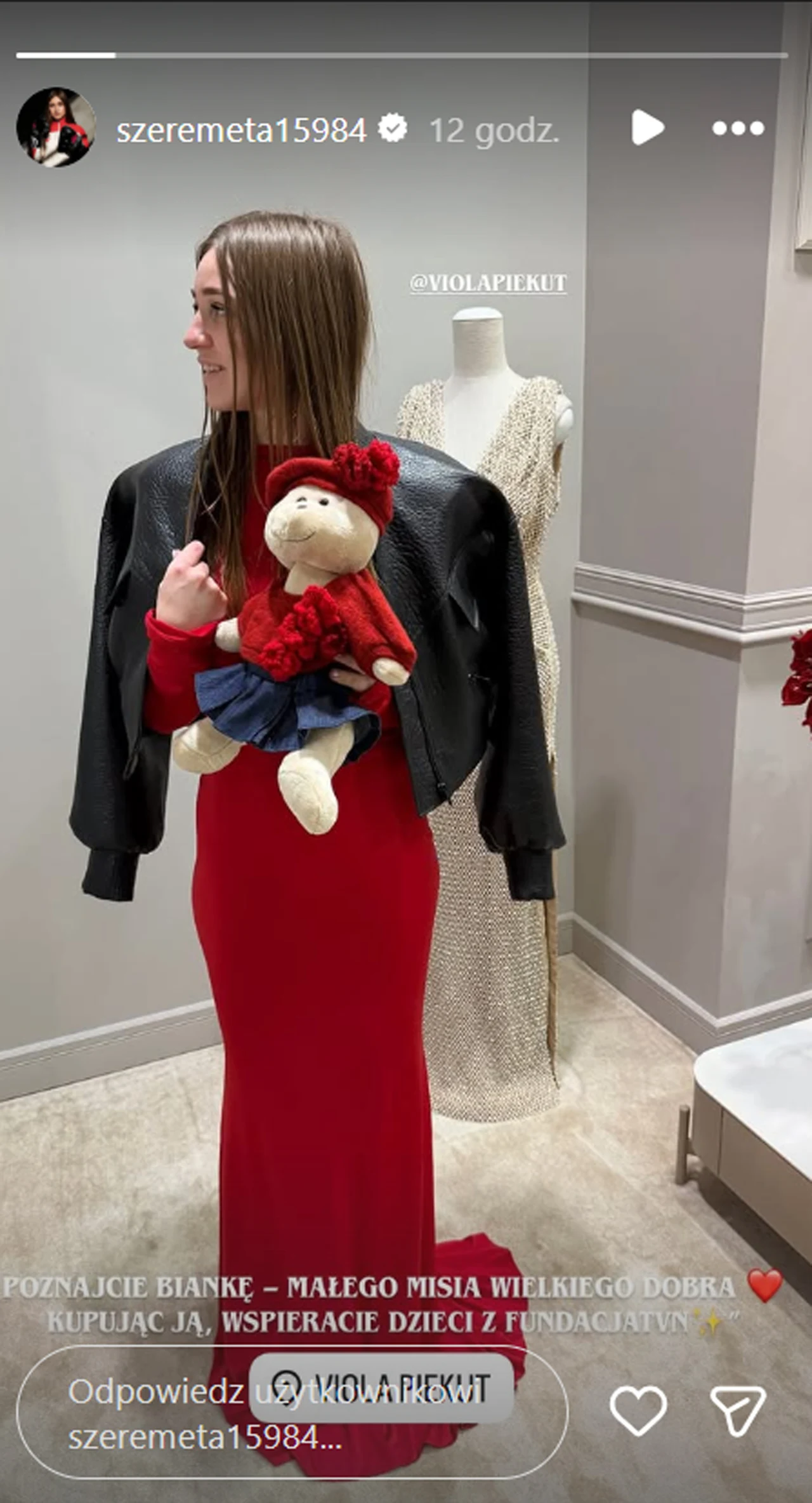 Julia Szeremeta Kobieta w długiej czerwonej sukni i czarnej kurtce narzuconej na ramiona trzyma maskotkę małej dziewczynki. W tle biały manekin ubrany w jasną suknię znajduje się blisko ściany w klimatycznym wnętrzu.