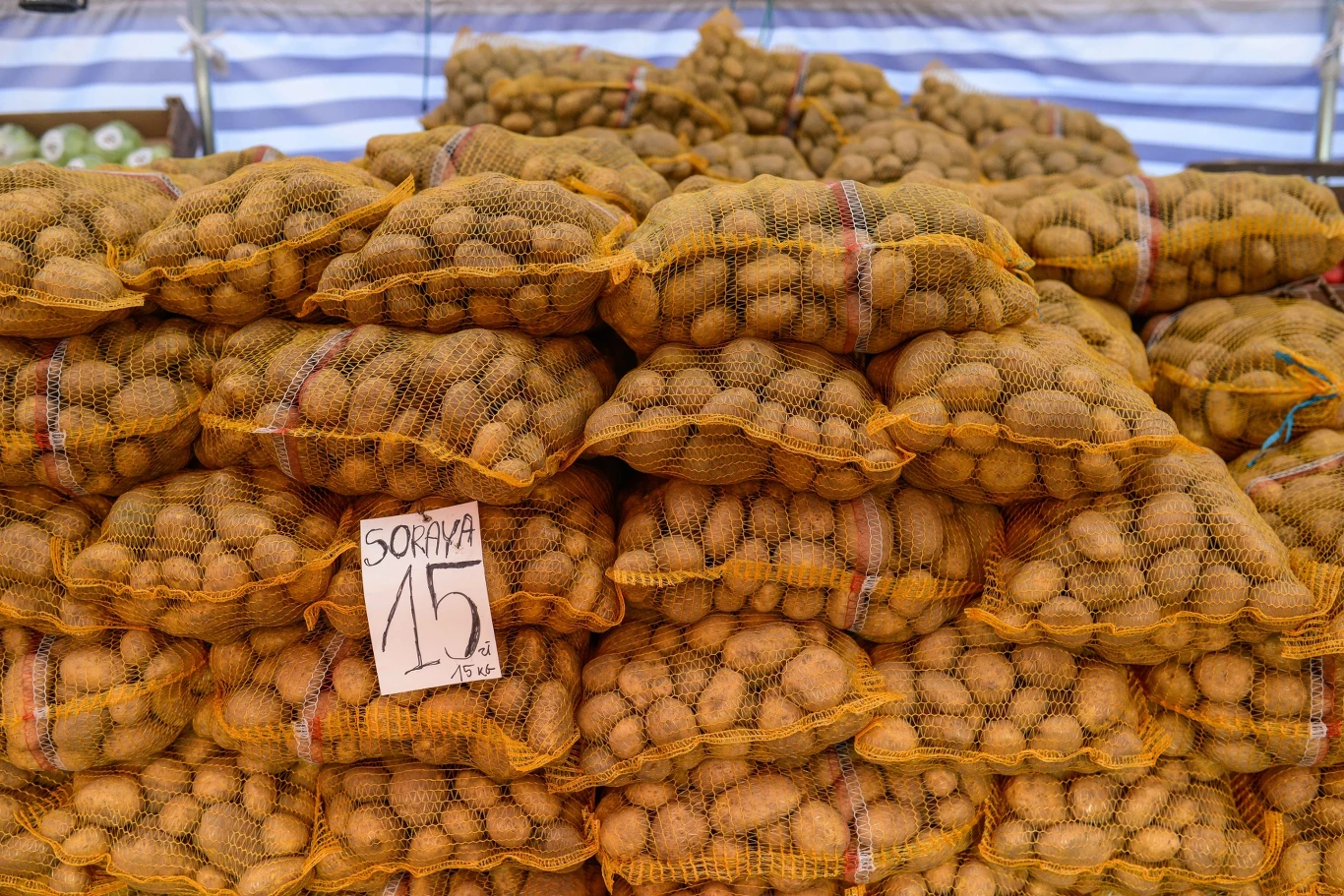 Polscy producenci mają problem ze sprzedażą ziemniaków. Mimo tego import rośnie. Stosy worków z ziemniakami ułożone na straganie, z widoczną ceną i nazwą odmiany na kartce papieru. W tle widać nieco rozmyte dodatkowe worki i straganowe otoczenie.