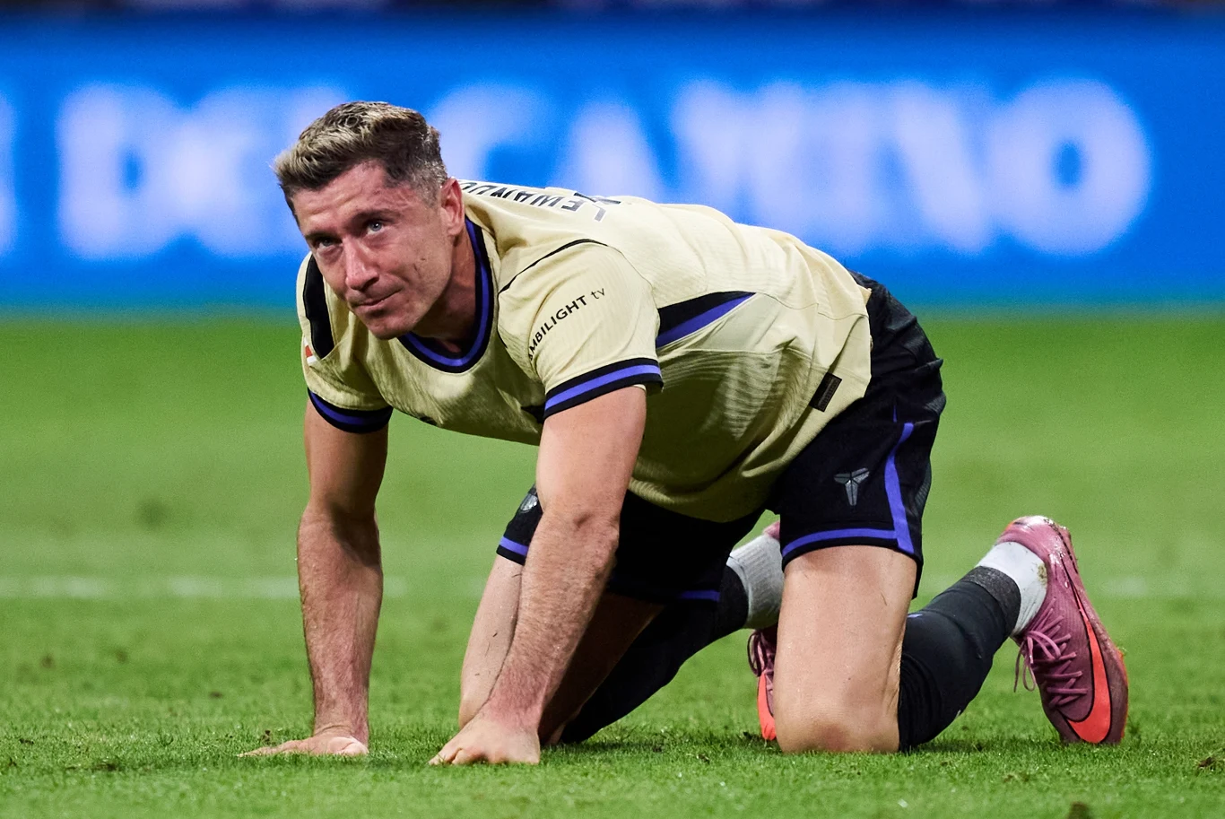 Robert Lewandowski Piłkarz ubrany w jasną koszulkę i ciemne spodenki klęczy na murawie boiska sportowego, z wyraźnie zmęczoną lub zawiedzioną miną, w tle widać rozmytą tablicę reklamową.