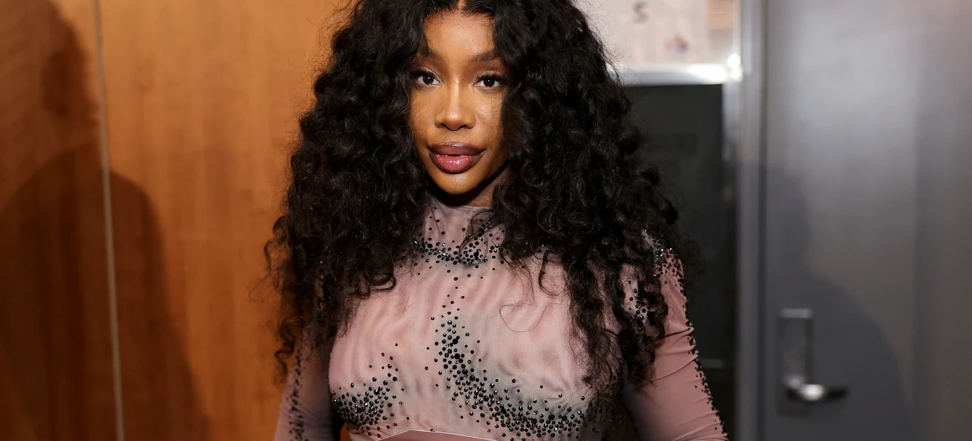 SZA Kobieta o długich, kręconych włosach ubrana w fioletową, prześwitującą bluzkę ozdobioną czarnymi kamieniami stoi na tle drewnianej ściany i szarych drzwi.