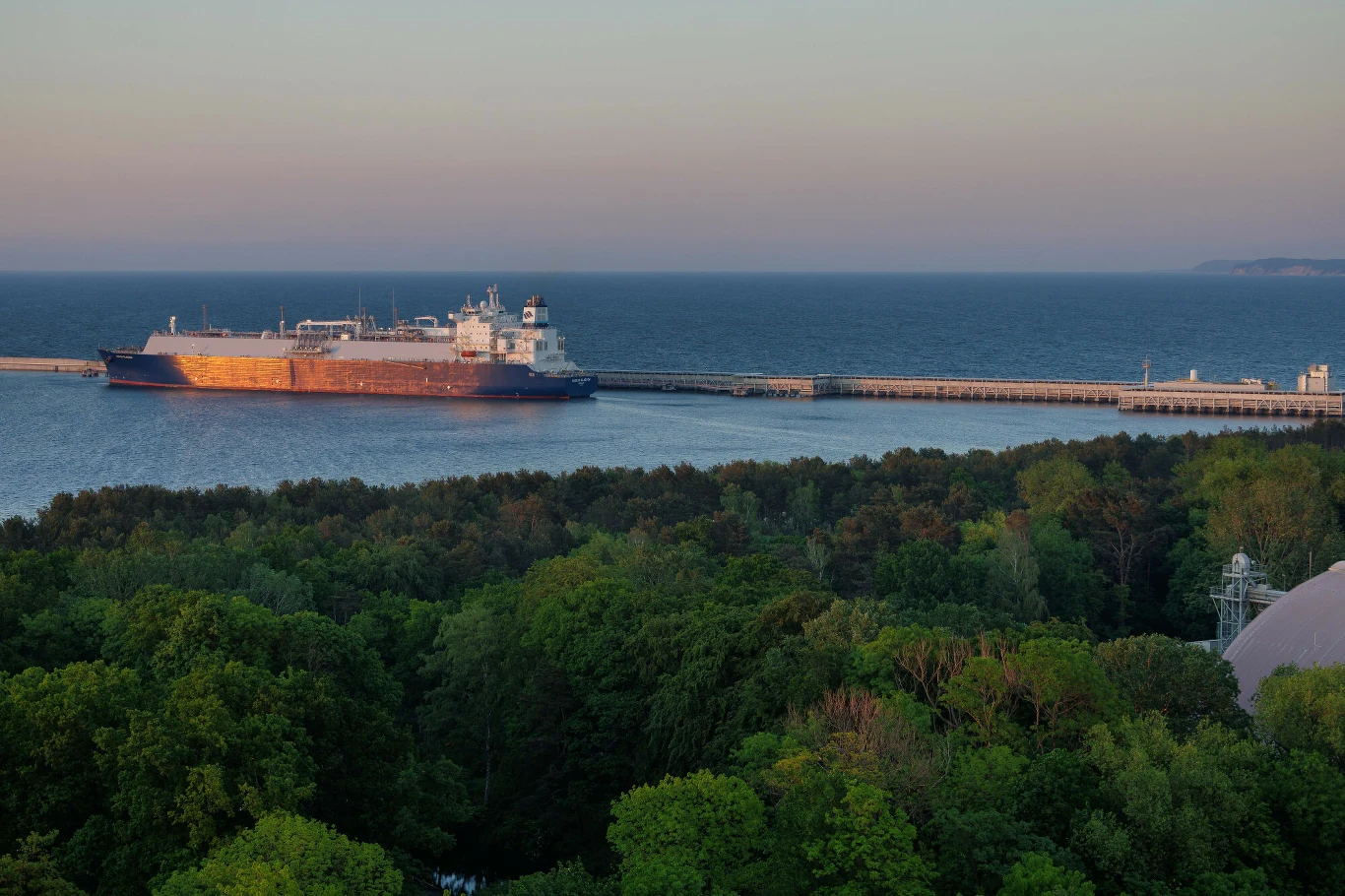 Gazoport zapewnia bezpieczeństwo energetyczne, ale turystyka odczuwa ograniczenia (zdj. ilustracyjne) Statek towarowy zacumowany przy długim nabrzeżu na tle spokojnego morza, w pierwszym planie gęsty, zielony las.