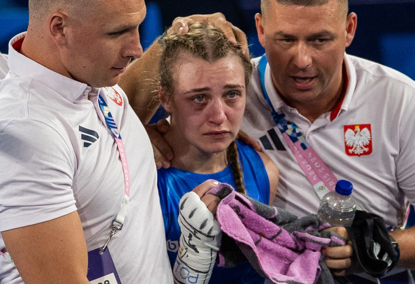 Julia Szeremeta ze swoim teamem Zawodniczka bokserska w niebieskim stroju siedzi pomiędzy dwoma trenerami. Wygląda na zmęczoną i wzruszoną, trzyma rękawicę oraz ręcznik. Trenerzy wspierają ją, jeden trzyma ją za głowę, drugi podaje wodę i ręcznik. Na ich koszulkach widnieje godło Pol...