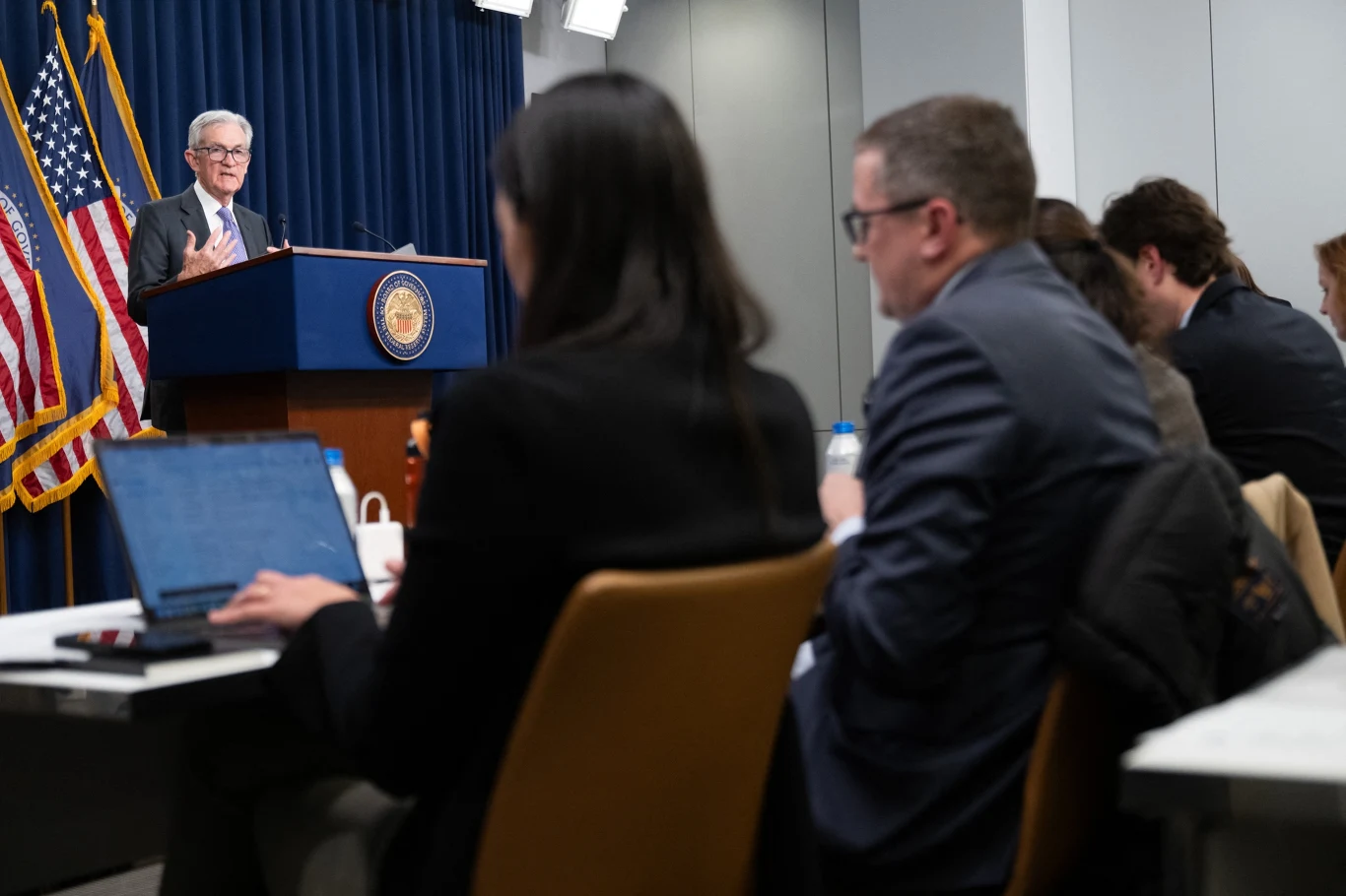 Grudniowa decyzja Rezerwy Federalnej o cięciu stóp nie została podjęta jednogłośnie. Konferencja prasowa z udziałem mężczyzny przemawiającego z mównicy z emblematem państwowym, za nim stoją flagi USA, przed sceną siedzą dziennikarze z laptopami i notatnikami.