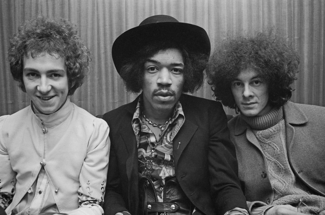 Noel Redding, Jimi Hendrix i Mitch Mitchell z The Jimi Hendrix Experience Trzech mężczyzn siedzi obok siebie. Centralna postać ma na sobie ciemny kapelusz i marynarkę, a z lewej i prawej strony znajdują się mężczyźni z charakterystycznie kręconymi fryzurami. Wszyscy patrzą bezpośrednio w obiektyw, a tło stanowi pionowo prążk...