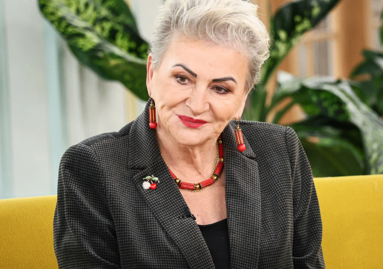 Hanna Bakuła Starsza kobieta z krótkimi, siwymi włosami siedzi na żółtej sofie. Ubrana jest w ciemny żakiet, pod nim czarny top. Ma na sobie czerwoną biżuterię: długie kolczyki i naszyjnik, a także broszkę w kształcie wiśni. W tle widoczne są zielone rośliny.