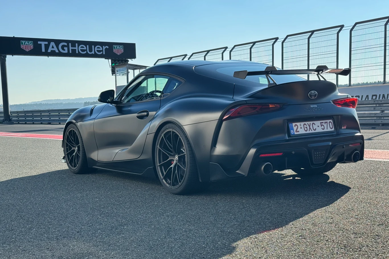 Czarny sportowy samochód Toyota Supra stoi na torze wyścigowym. W tle widać ogrodzenie i tablicę reklamową TAG Heuer. Auto zwrócone tyłem, widoczne szerokie nadkola, duży spoiler na klapie bagażnika oraz sportowe felgi.