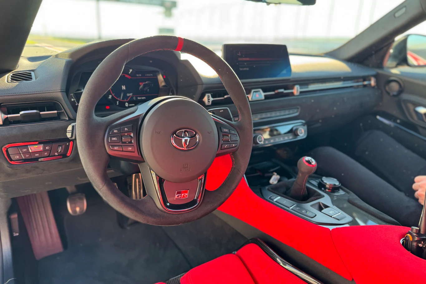 Wnętrze sportowego samochodu marki Toyota z widocznym logotypem na kierownicy, nowoczesną deską rozdzielczą, dużym ekranem multimedialnym oraz czerwonym wykończeniem siedzeń.