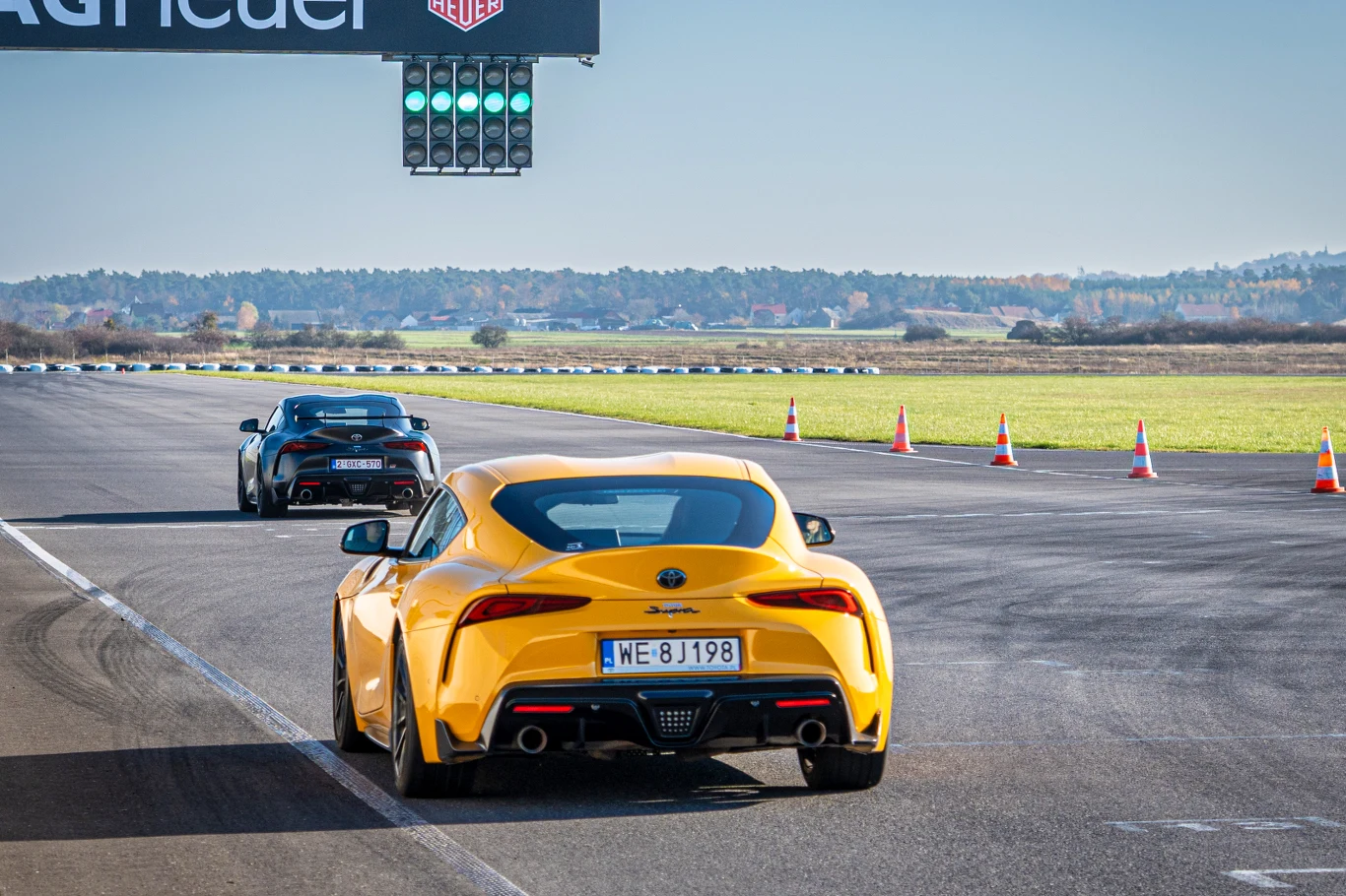 Żółty sportowy samochód marki Toyota Supra na torze wyścigowym, za nim jedzie czarny samochód tej samej marki. W tle widoczny sygnalizator świetlny oraz barierki odgradzające trasę. Otoczenie stanowi otwarty, zielony teren poza miastem.