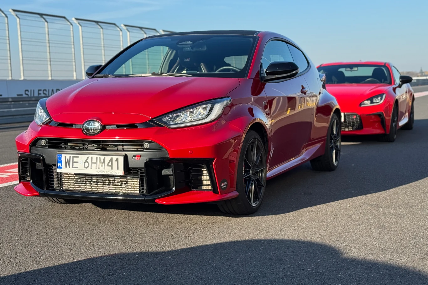 Dwa czerwone sportowe samochody marki Toyota ustawione jeden za drugim na torze wyścigowym, na pierwszym planie Toyota GR Yaris z widoczną tablicą rejestracyjną, w tle szeroka asfaltowa droga oraz barierki toru.