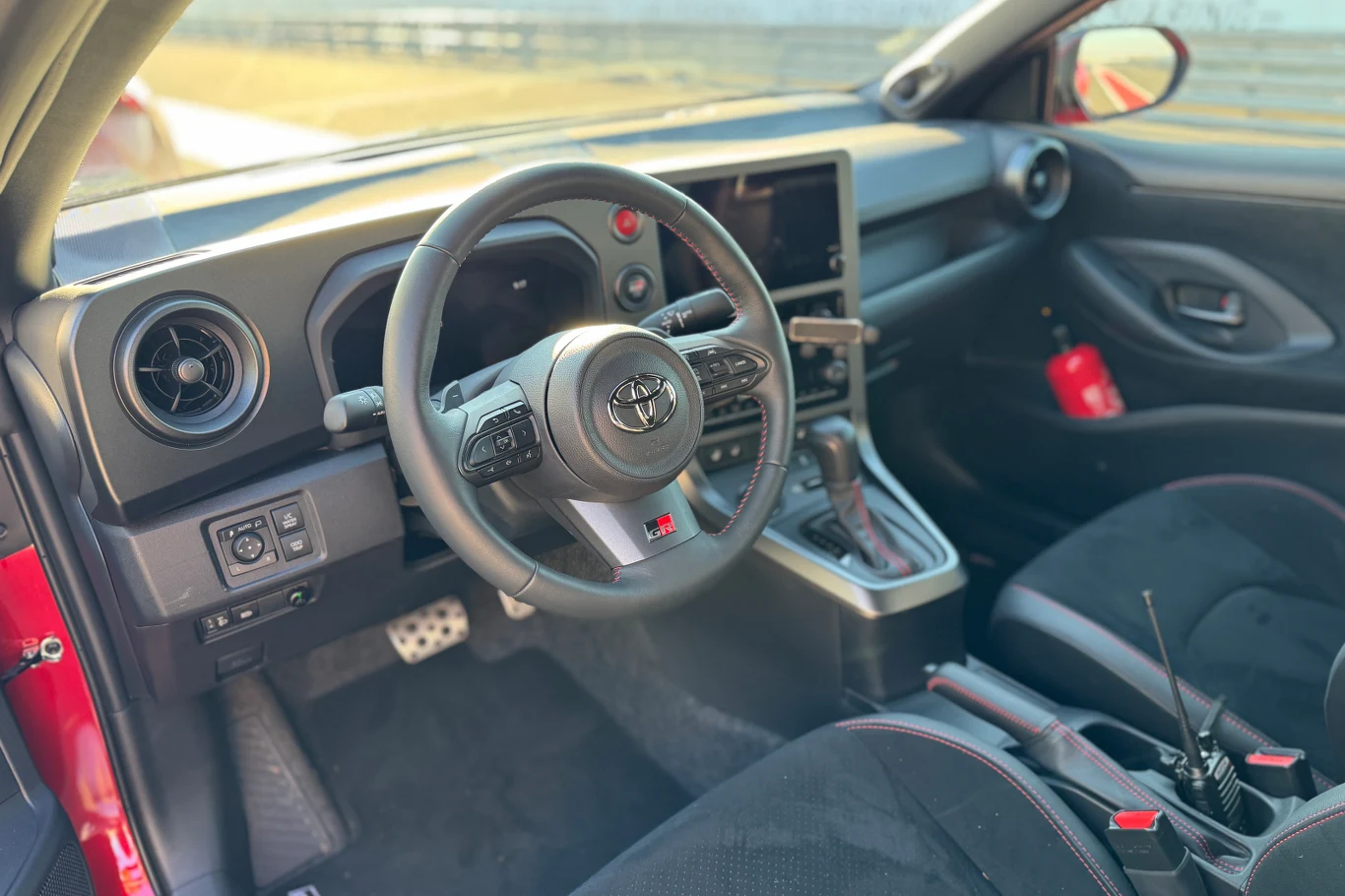 Kierownica z logo Toyoty, cyfrowy wyświetlacz, centralna konsola z dźwignią zmiany biegów i nowoczesnym panelem sterowania, czarne siedzenia z czerwonymi przeszyciami oraz radiostacja na siedzeniu pasażera.