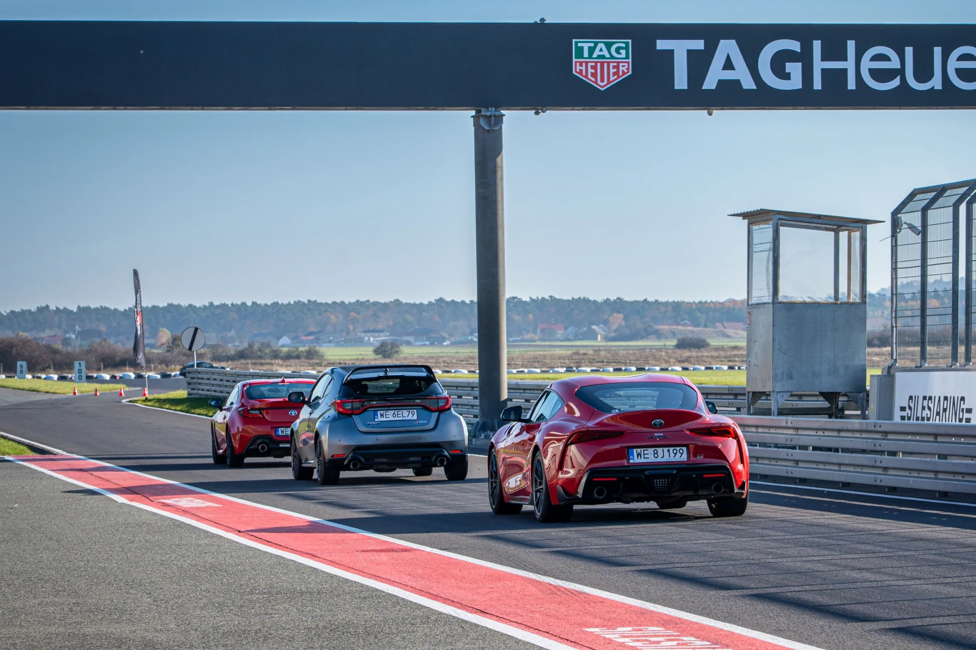 Trzy sportowe samochody, w tym czerwony i niebieski, poruszają się po torze wyścigowym pod bramką reklamową TAG Heuer na tle otwartej przestrzeni i trybuny