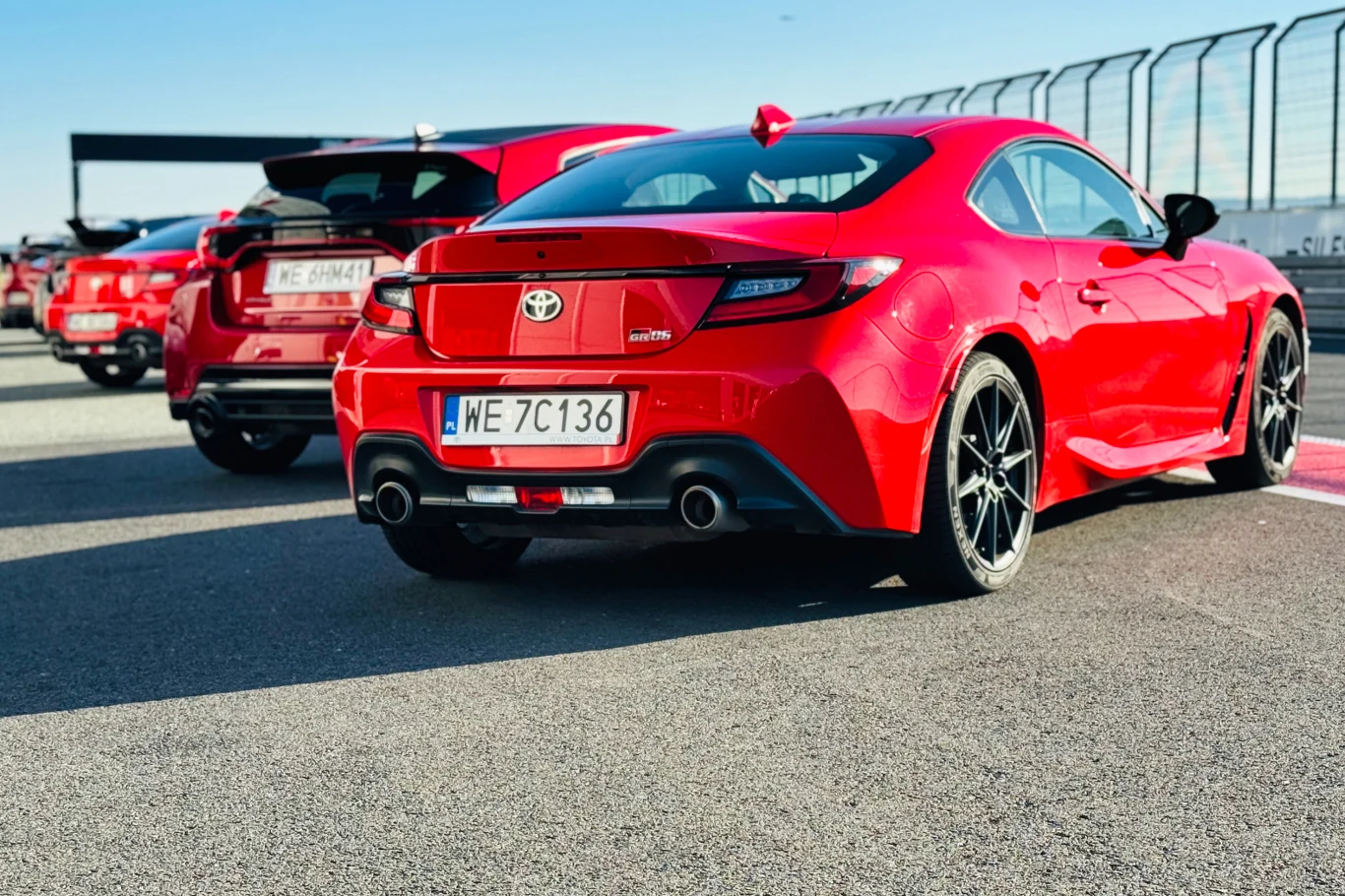 Czerwone sportowe samochody Toyota GR86 ustawione na torze wyścigowym, w tle widoczny fragment barier ochronnych i inne auta gotowe do startu.