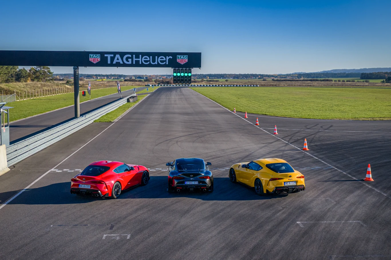 Trzy sportowe samochody stoją obok siebie na torze wyścigowym w słoneczny dzień, widoczne są także pachołki wyznaczające trasę oraz banner TAG Heuer nad linią mety.