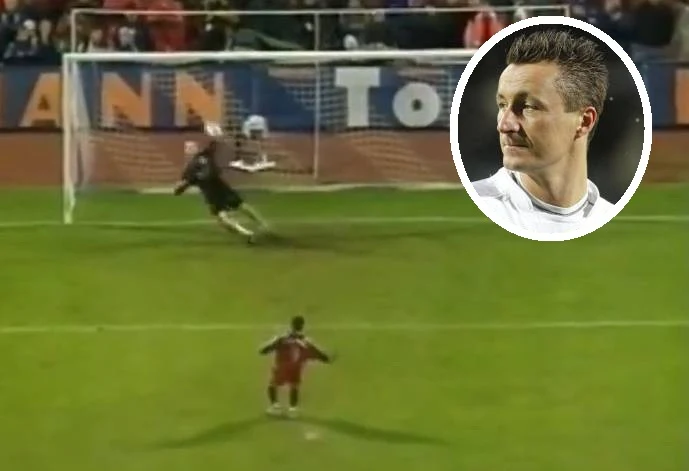 Mirosław Dreszer zatrzymał Bayern Monachium. Potem na drodze stanął Tomasz Hajto Piłkarz wykonuje rzut karny na stadionie podczas meczu, bramkarz rzuca się w stronę piłki, a w prawym górnym rogu umieszczono portret mężczyzny w białej koszulce, zwróconego w bok.