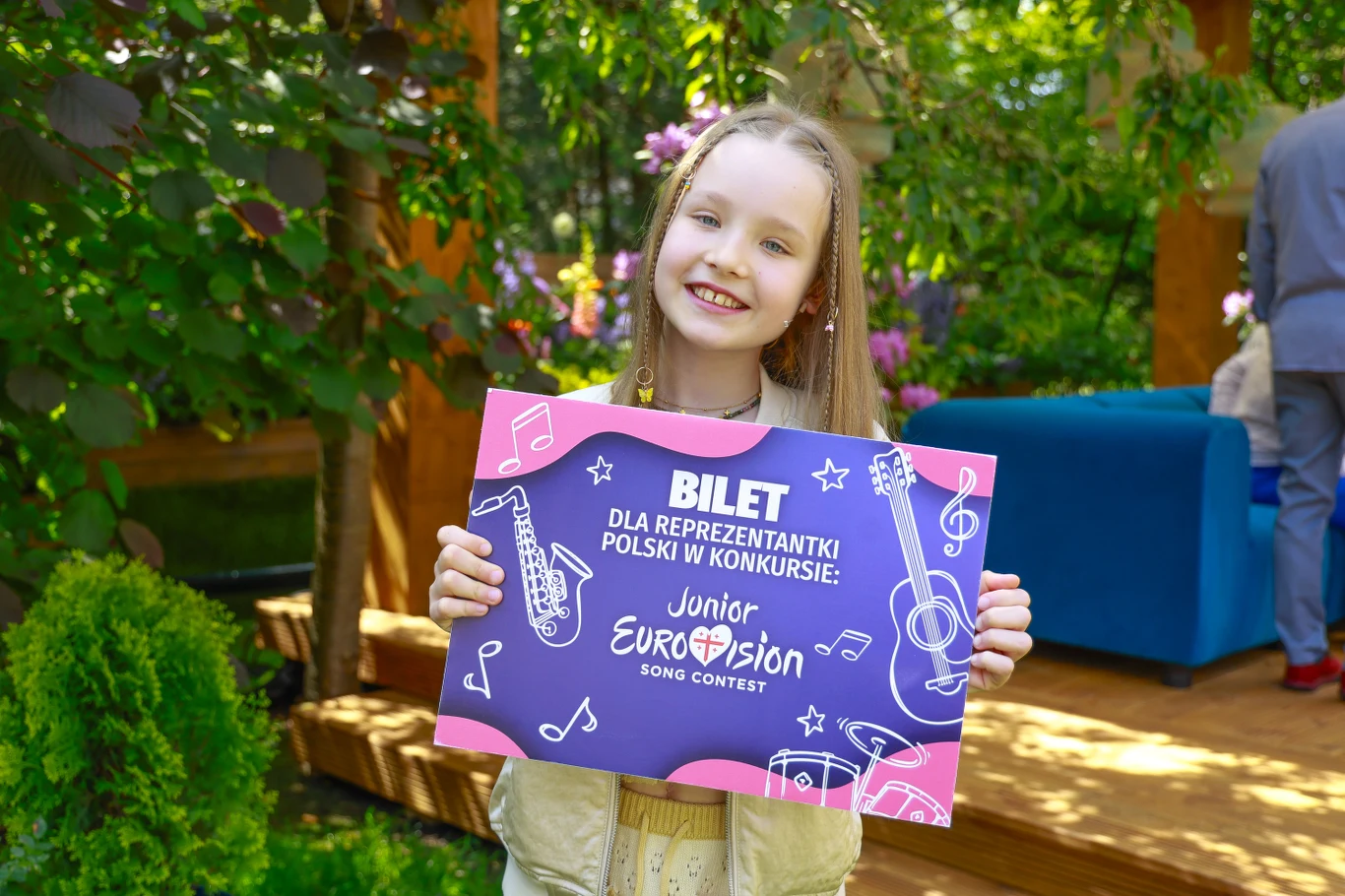 Marianna Kłos trenuje przed występem na Eurowizji Junior 2025 Młoda dziewczynka z długimi blond włosami trzyma w rękach kolorowy plakat informujący o bilecie dla reprezentantki Polski w konkursie Junior Eurovision Song Contest, stojąc w ogrodzie pełnym zieleni i kwiatów.
