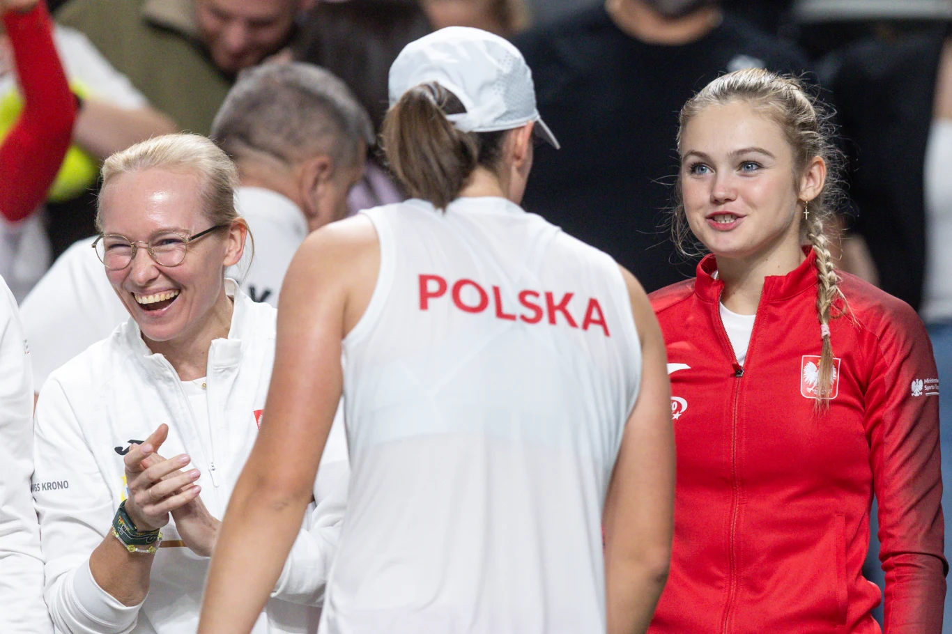 Trzy kobiety w sportowych strojach z emblematami Polski podczas rozmowy i radosnej wymiany uśmiechów na tle innych osób, centralna postać w czapce i koszulce z napisem POLSKA odwrócona plecami.