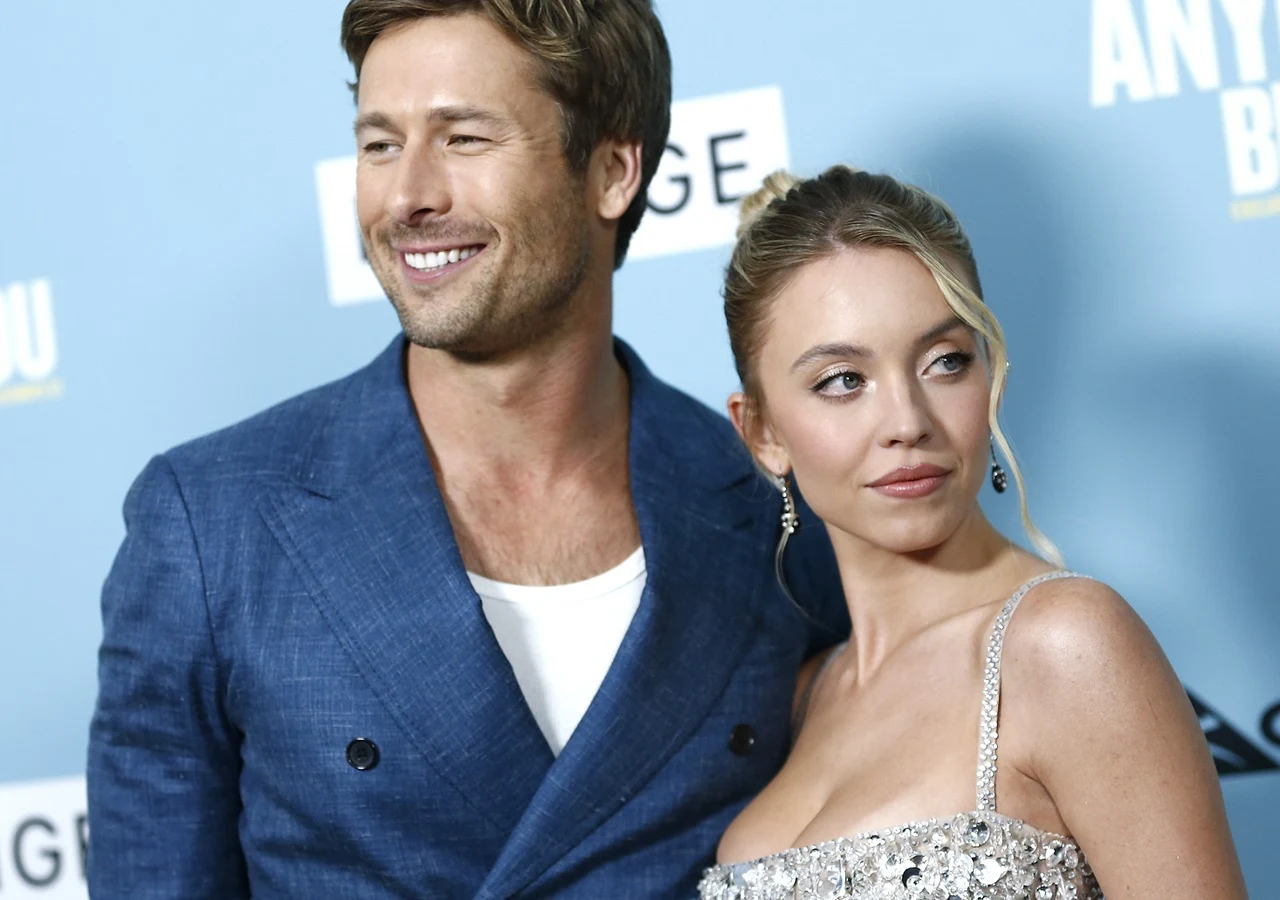 Glen Powell, Sydney Sweeney Dwójka elegancko ubranych osób pozuje razem na tle niebieskiej ściany z napisem, mężczyzna uśmiecha się i obejmuje kobietę, która ma upięte włosy i błyszczącą sukienkę na ramiączkach.