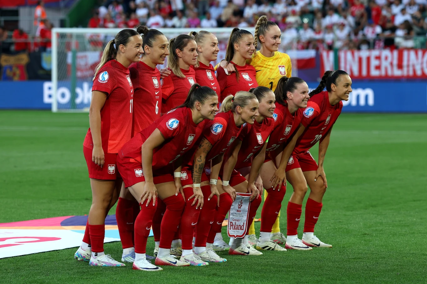 Kobieca reprezentacja Polski Grupa zawodniczek polskiej reprezentacji piłkarskiej w czerwonych strojach stoi w rzędzie na murawie stadionu przed rozpoczęciem meczu, w tle widownia z kibicami i biało-czerwonymi flagami.