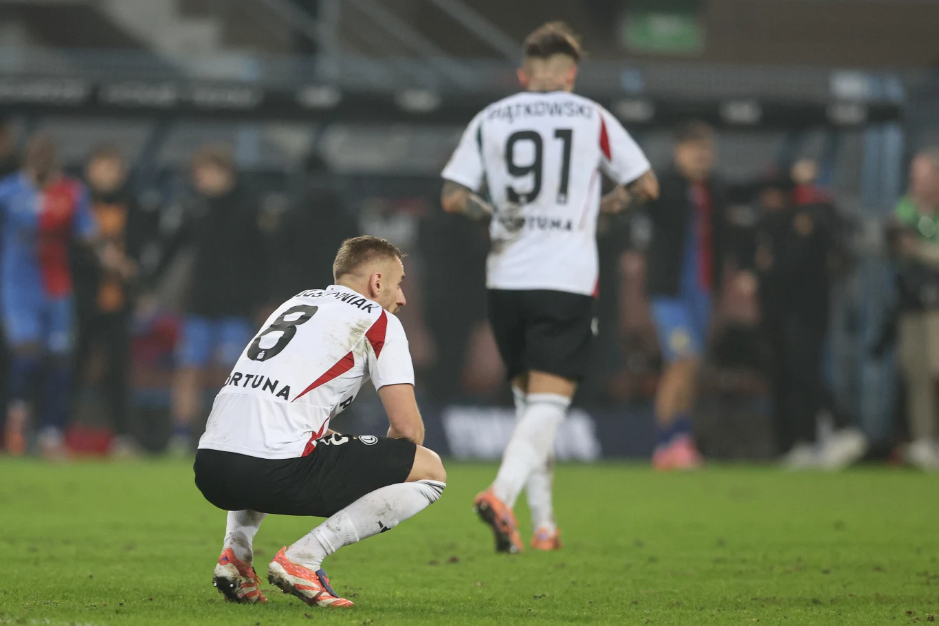 Legia Warszawa Dwóch piłkarzy w białych koszulkach znajduje się na murawie, jeden z nich kuca, drugi odchodzi z widocznym na plecach numerem 91. W tle rozmyte postacie i trybuny stadionu.