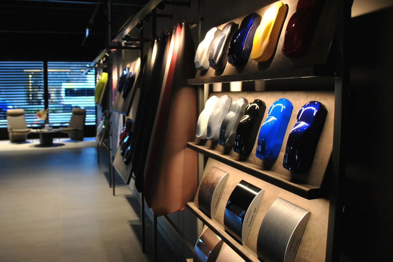 Nowy projekt ma pozwalać na budowanie wyjątkowych samochodów Kolorowe próbki lakierów samochodowych zawieszone na półkach, obok nich naświetlone tkaniny i próbki tapicerek wnętrz, a w tle widoczne są nowoczesne fotele i duże okno wpuszczające światło.