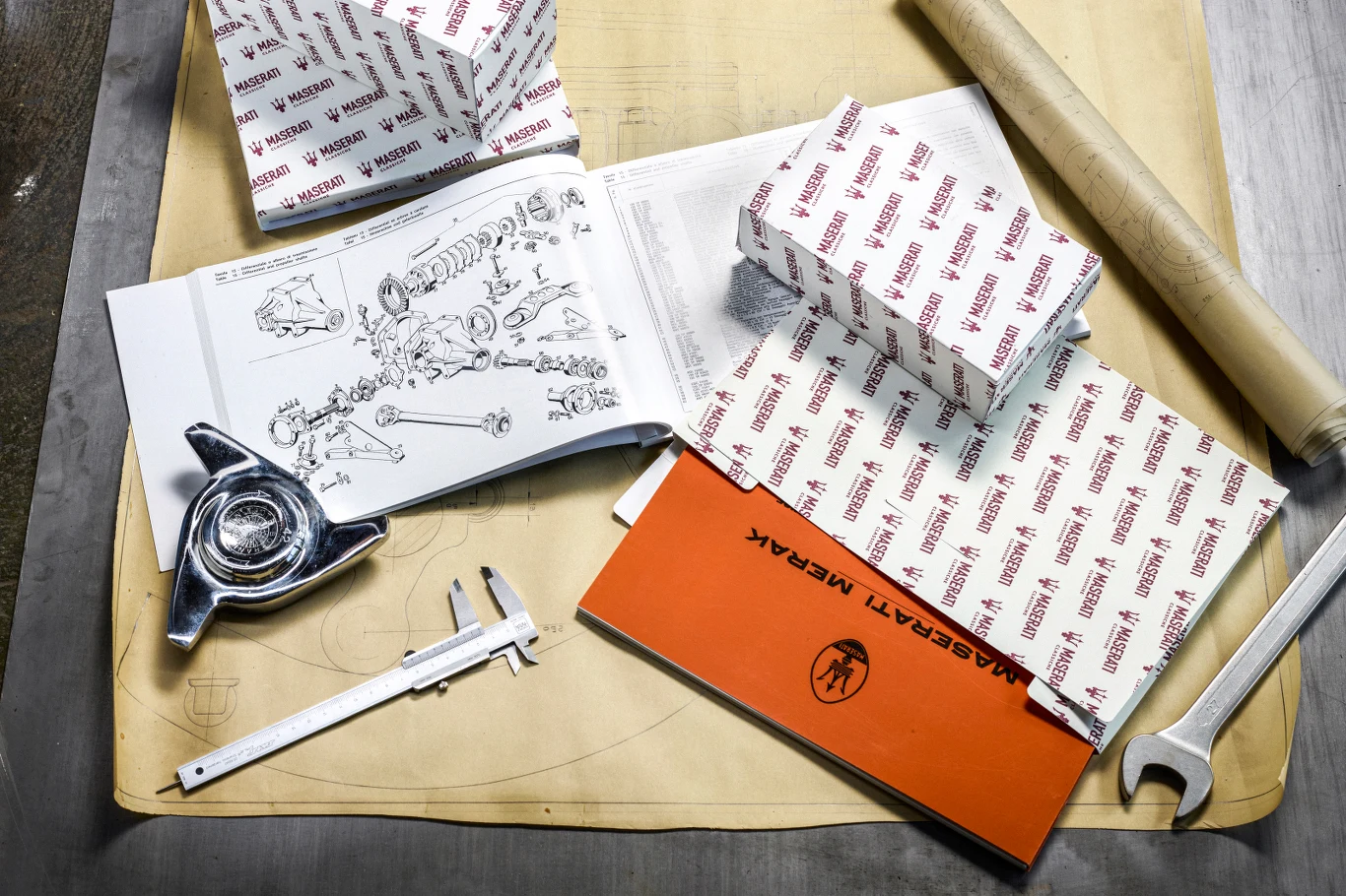 Klienci Alfy Romeo i Maserati będą mogli w większym stopniu indywidualizować swoje auta Elementy techniczne związane z motoryzacją, w tym części Maserati zapakowane w firmowe pudełka, otwarty katalog z diagramem technicznym części samochodowych, suwmiarka, klucz i zwinięty plan, wszystko rozłożone na blacie roboczym.