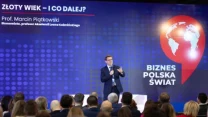 Mężczyzna w garniturze przemawia do publiczności na tle dużego ekranu z napisem 'Złoty Wiek – I co dalej?' oraz grafiką globu i napisem 'Biznes Polska Świat'. Publiczność z uwagą słucha wykładu.