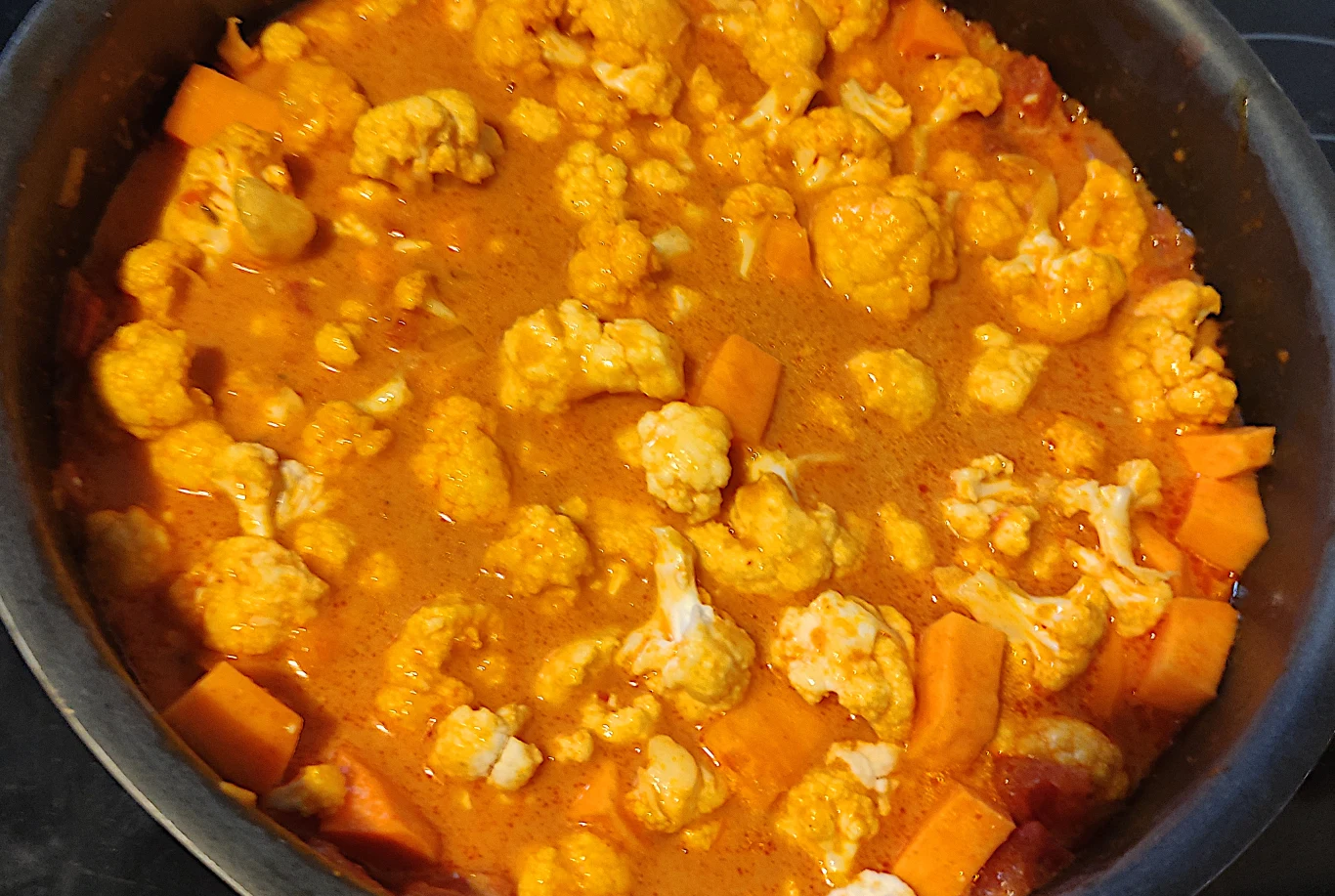 Kawałki kalafiora i batatów w sosie curry w garnku, pomarańczowa barwa potrawy podkreślona przez przyprawy i kremową konsystencję.