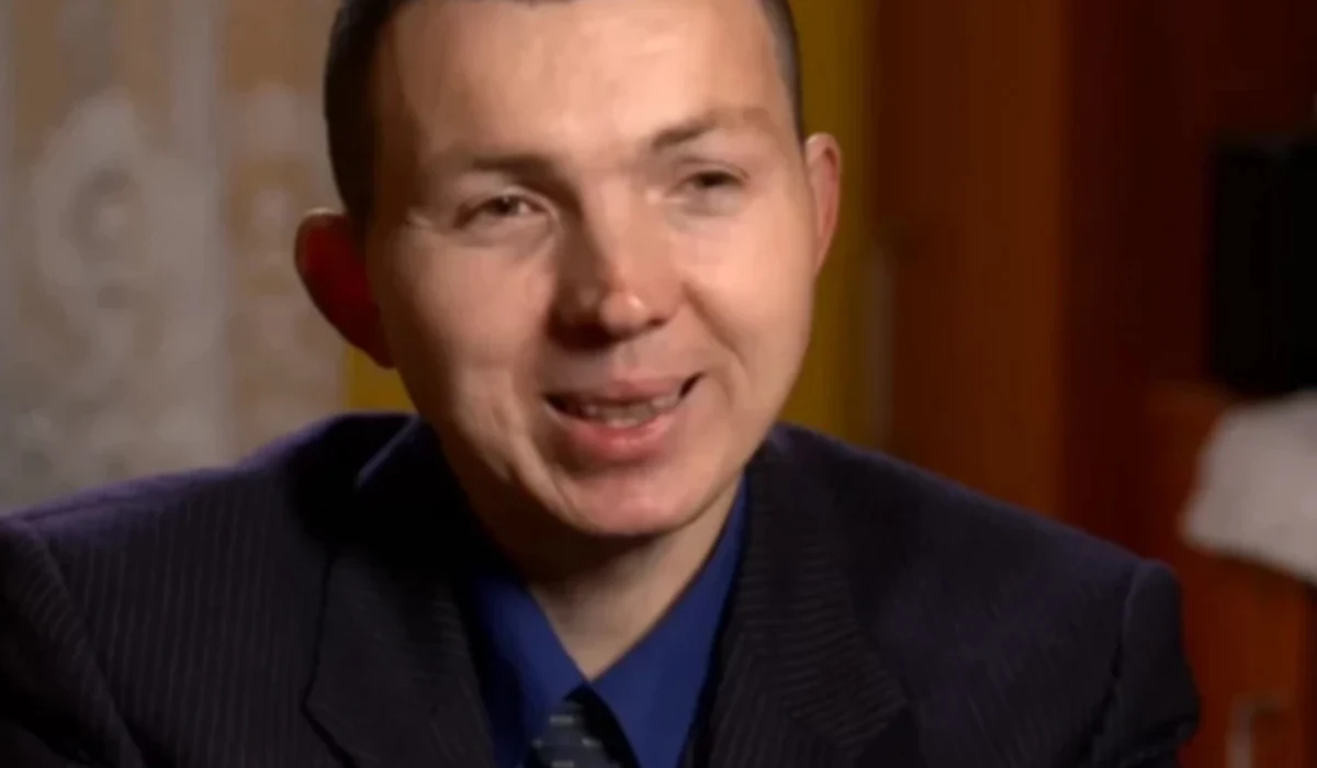 Jaruś z "Chłopaków do wzięcia" Mężczyzna w garniturze, uśmiechający się, siedzi w pomieszczeniu o ciepłym oświetleniu. W tle rozmyte elementy dekoracyjne i drewniane meble.