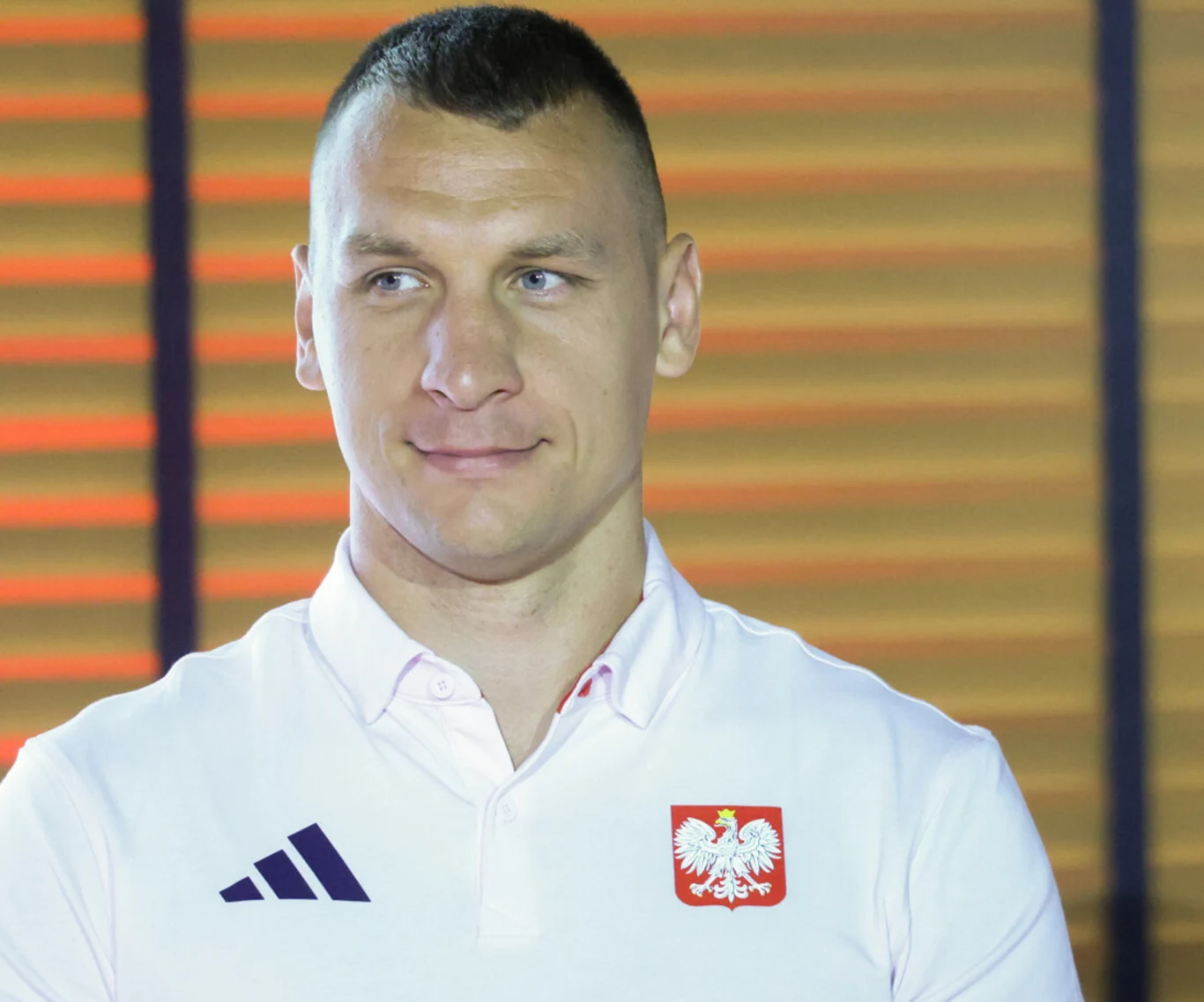 Mężczyzna w białej koszulce polo z godłem Polski i logo sportowej marki stoi na tle poziomych pomarańczowych żaluzji, patrząc w bok z delikatnym uśmiechem