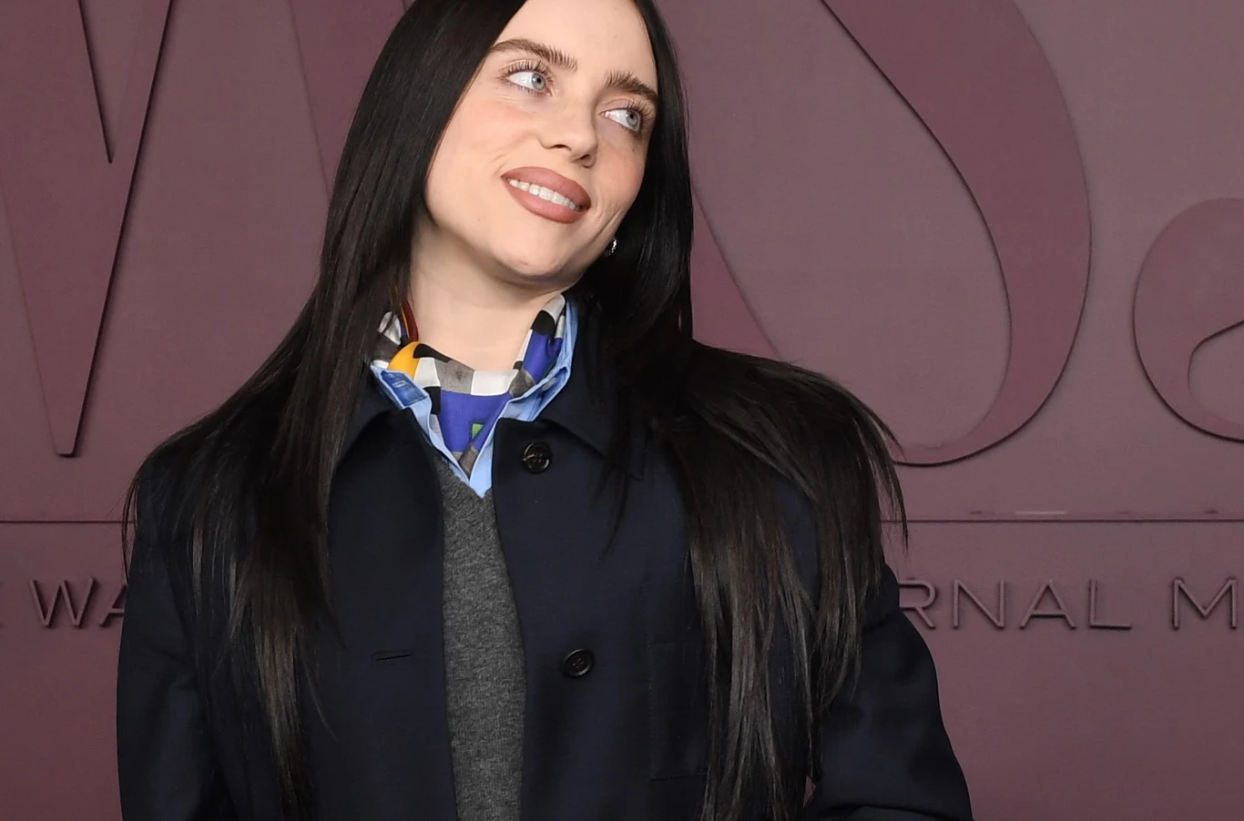 Billie Eilish współpracowała z Jamesem Cameronem przy tworzeniu filmu trasy Młoda kobieta z długimi, ciemnymi włosami pozuje na tle ściany z tłoczonymi literami, ubrana w czarny płaszcz, szary sweter i kolorową apaszkę, uśmiecha się delikatnie patrząc w bok.