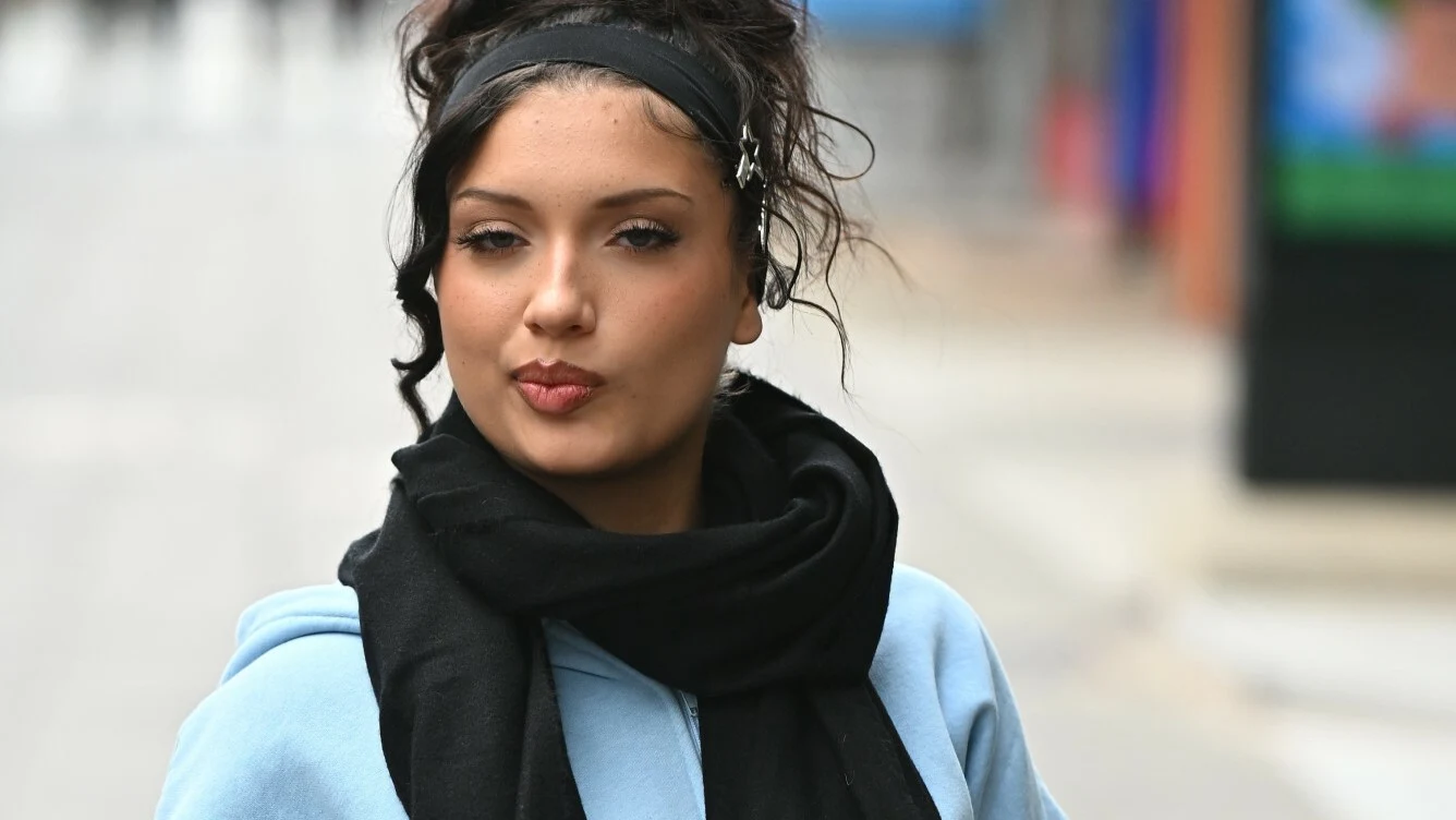 Viki Gabor Młoda kobieta z ciemnymi włosami spiętymi opaską, ubrana w jasnoniebieską bluzę i czarny szalik, spogląda lekko w bok na tle rozmytej ulicy miejskiej.