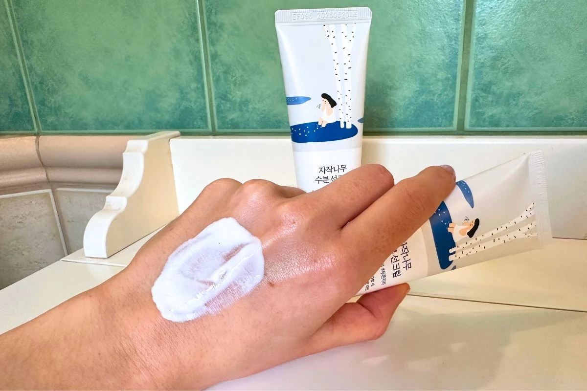 Dłoń z nałożonym kremem na wierzchu, w tle dwie tubki kosmetyku z ilustracją siedzącej kobiety i napisami w języku koreańskim na opakowaniach.