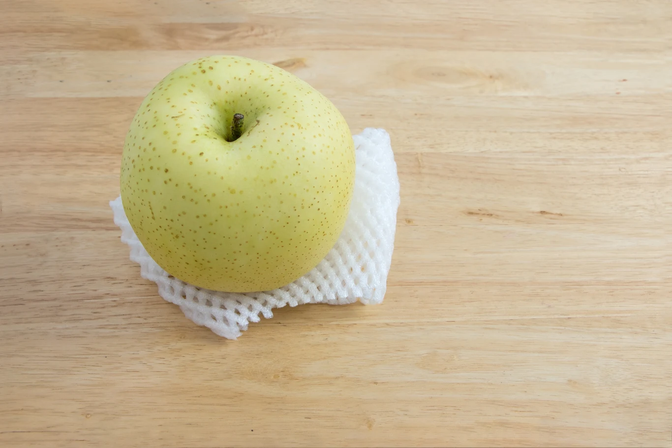Jabłko o żółtej skórce z delikatnymi kropeczkami ułożone na białej piance ochronnej, leżące na jasnym, drewnianym blacie.