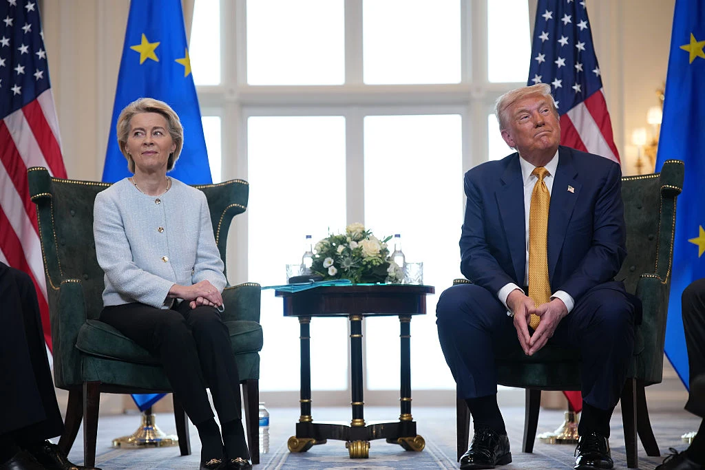 Szefowa Komisji Europejskiej Ursula von der Leyen i prezydent USA Donald Trump Dwoje polityków siedzi na fotelach naprzeciw siebie przy małym stoliku z kwiatami, w tle widoczne flagi Unii Europejskiej oraz Stanów Zjednoczonych.