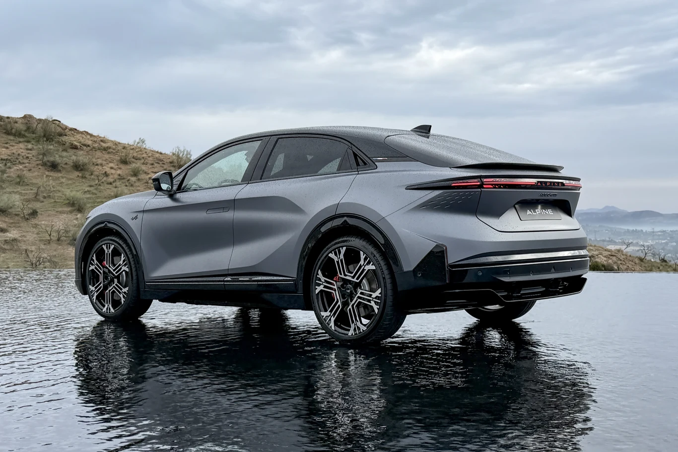 Nowoczesny, grafitowy samochód elektryczny typu SUV stoi na lśniącej nawierzchni, odbijając się w wodzie. W tle lekko pochmurne niebo i delikatnie pagórkowate tereny.