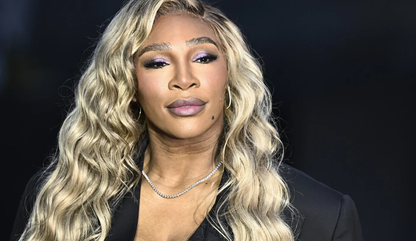 Serena Williams Kobieta o długich, falowanych blond włosach z delikatnym makijażem, ubrana w czarny strój i srebrny łańcuszek, patrzy spokojnie w stronę aparatu. Tło jest ciemne, co eksponuje jej twarz i fryzurę.