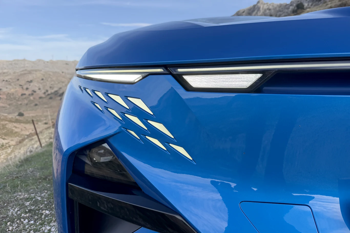 Fragment przedniego zderzaka i reflektora nowoczesnego samochodu w intensywnym niebieskim kolorze z charakterystycznymi, geometrycznymi wstawkami świetlnymi.