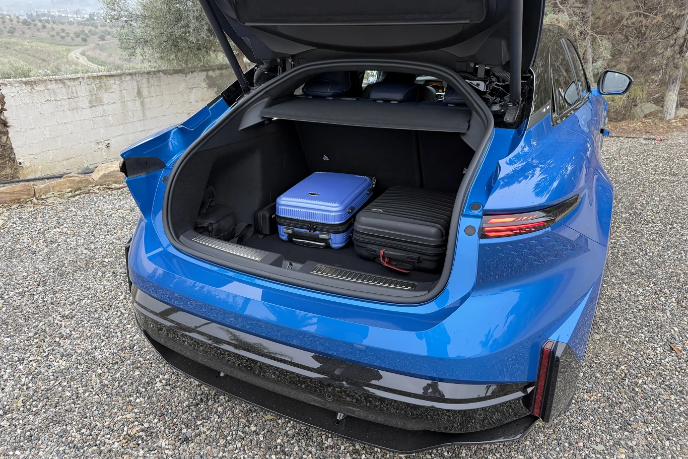 Otwarty bagażnik niebieskiego samochodu zawiera trzy walizki, w tym jedną niebieską i dwie czarne, ustawione na żwirowym podłożu, w tle widoczne są drzewa i niski mur.