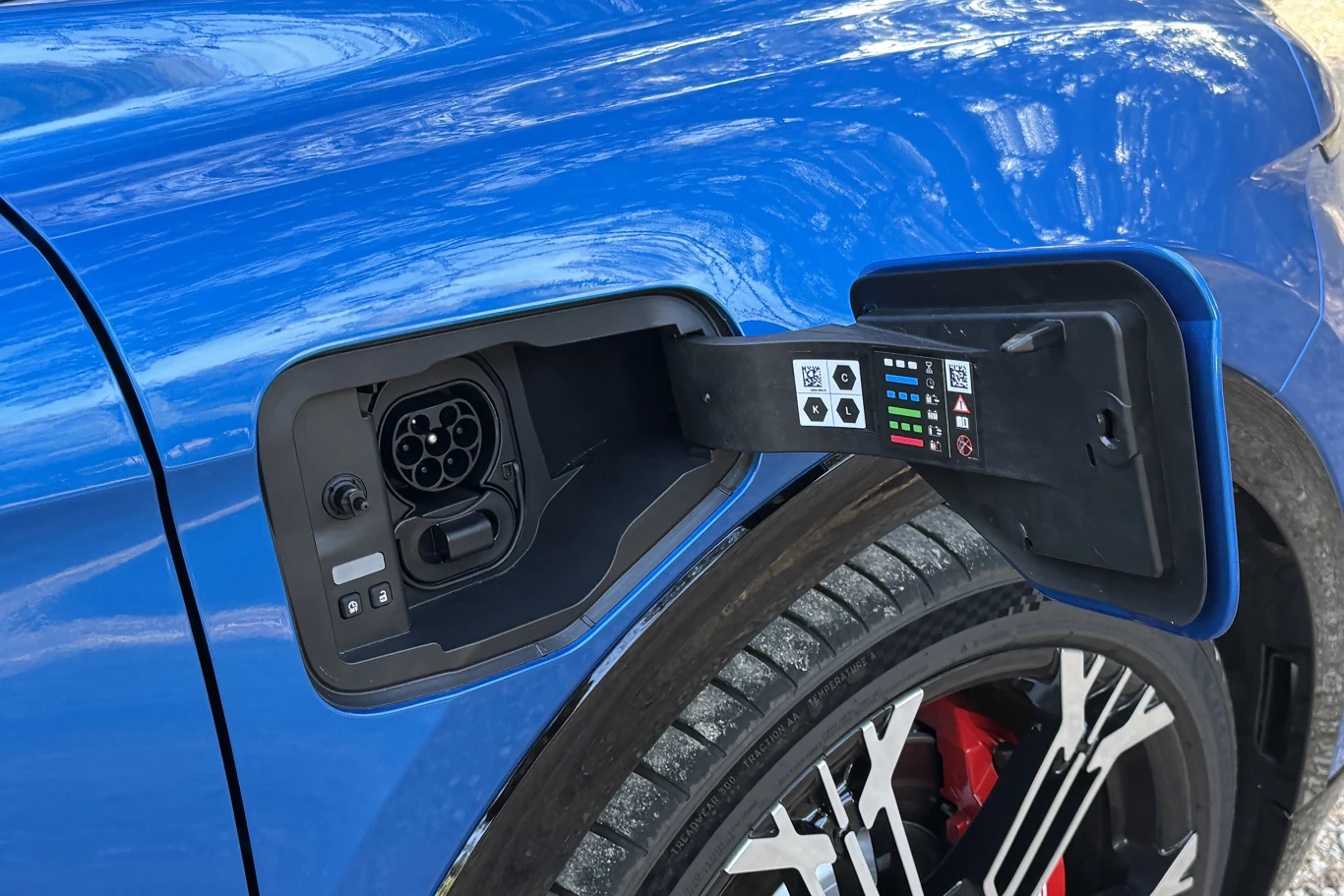 Otwarty port ładowania samochodu elektrycznego w niebieskim pojeździe, widoczne szczegółowe oznaczenia przy złączu i fragment felgi z czerwonym zaciskiem hamulcowym.